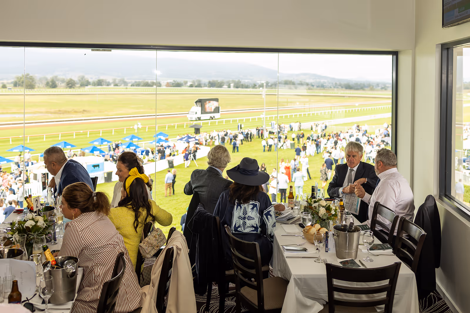 People seated at a table with drinks and flowers in a room overlooking a horse racing track with crowds and tents outside.