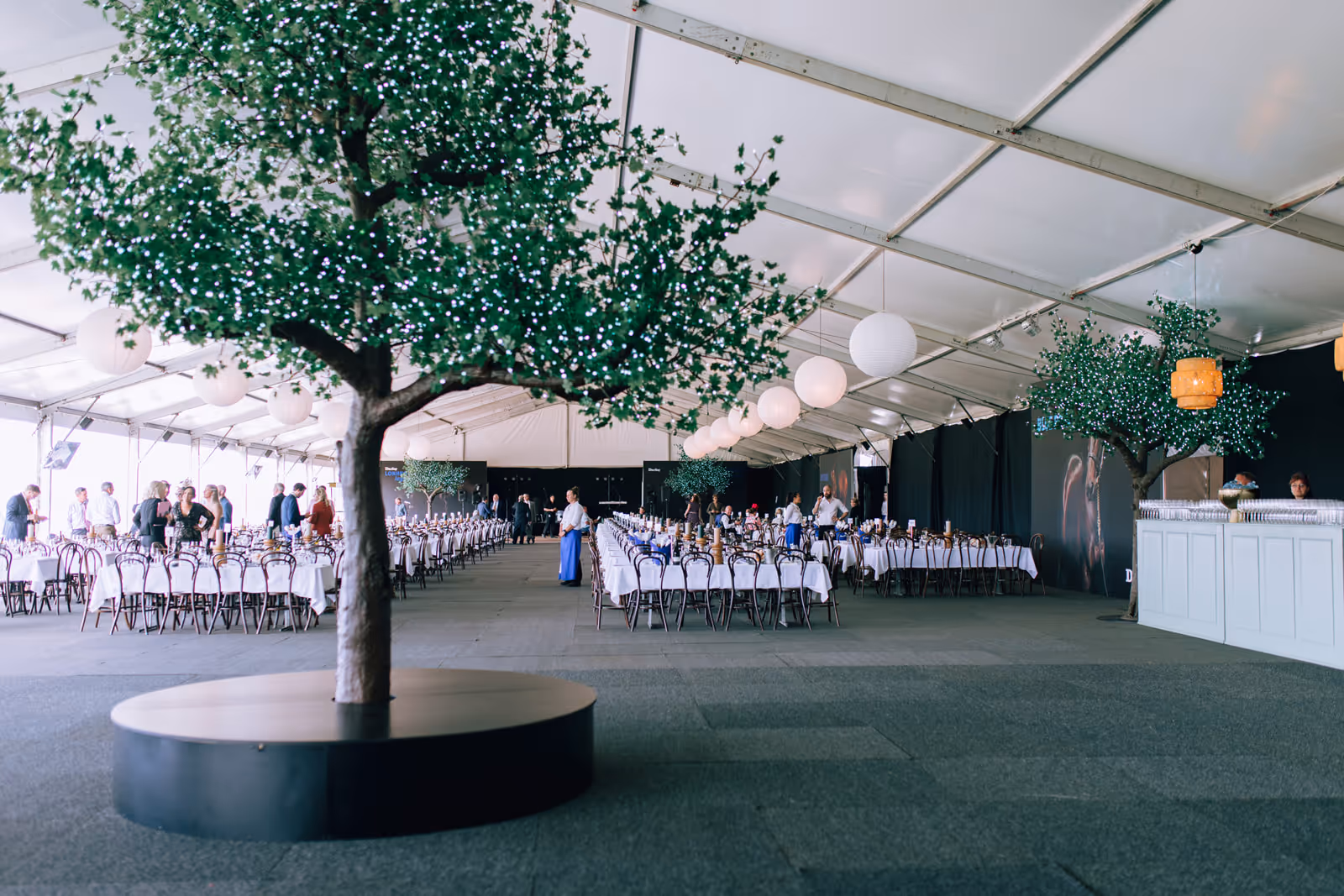 Large white event marquee with long tables set for dining, decorated with artificial trees adorned with small white lights and hanging paper lanterns.