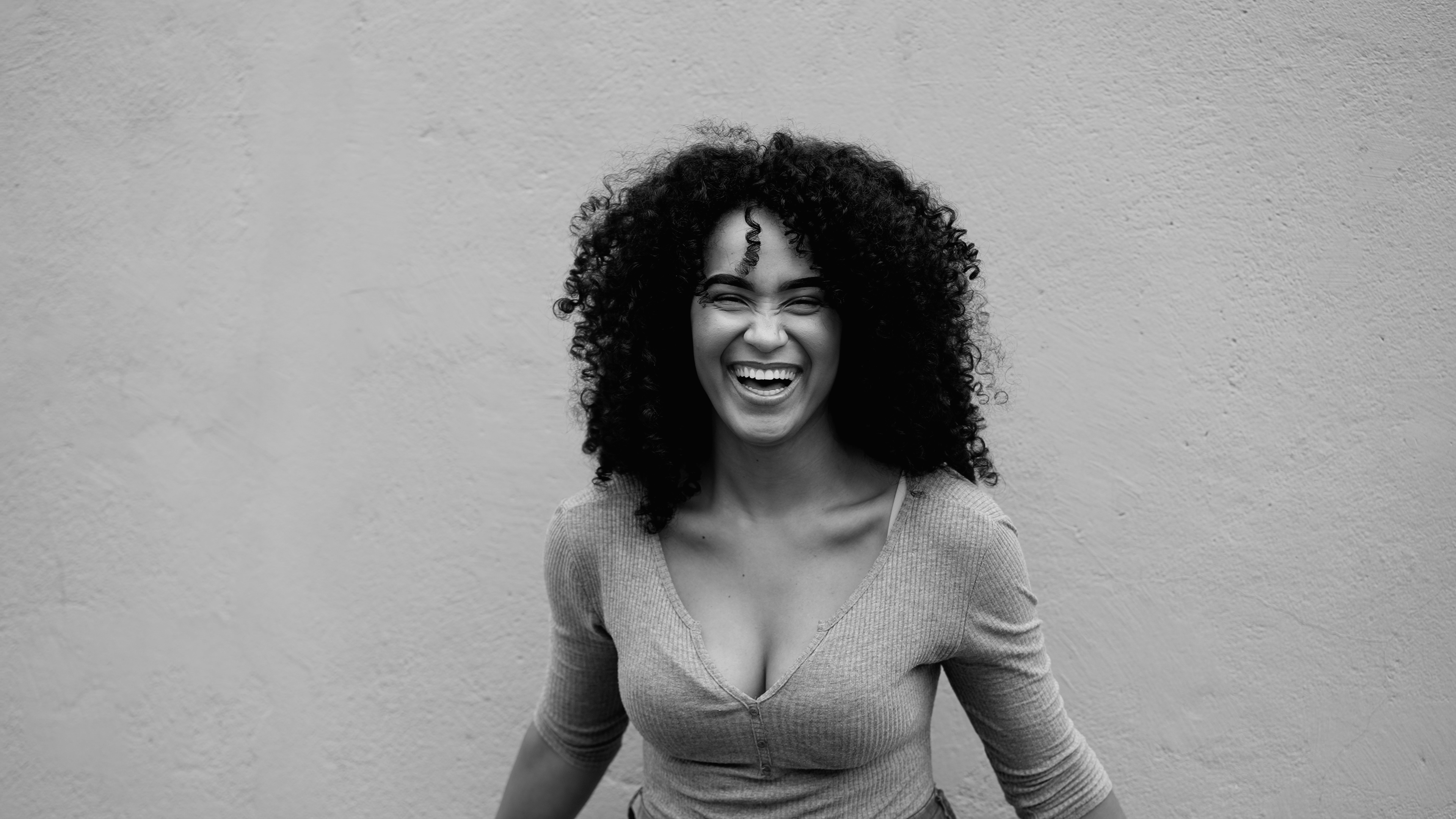 Smiling woman with curly hair wearing a ribbed V-neck top against a textured wall.