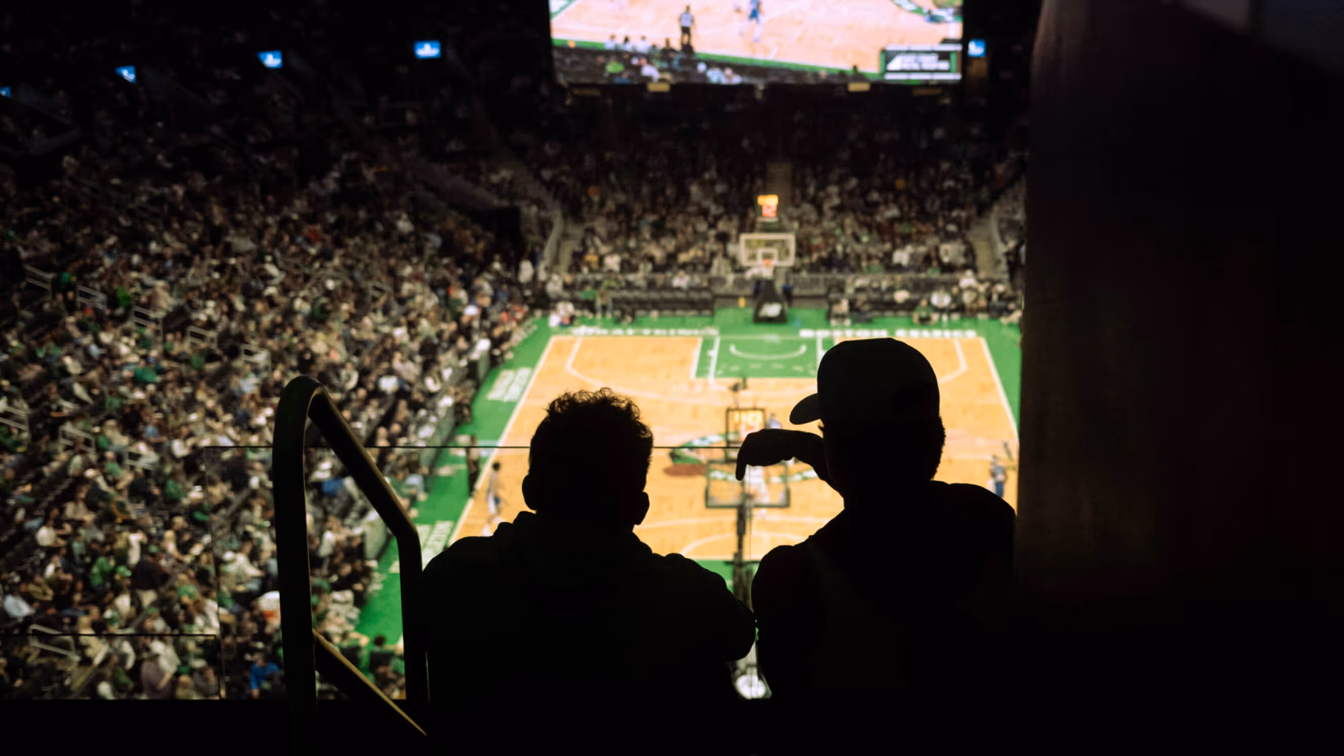 Two silhouetted spectators sitting in a stadium overlooking a basketball court during a game.