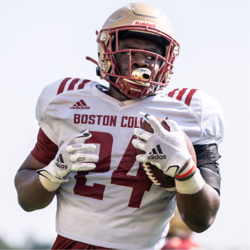 Boston College football player wearing number 24 jersey, gold helmet, and white gloves holding a football.