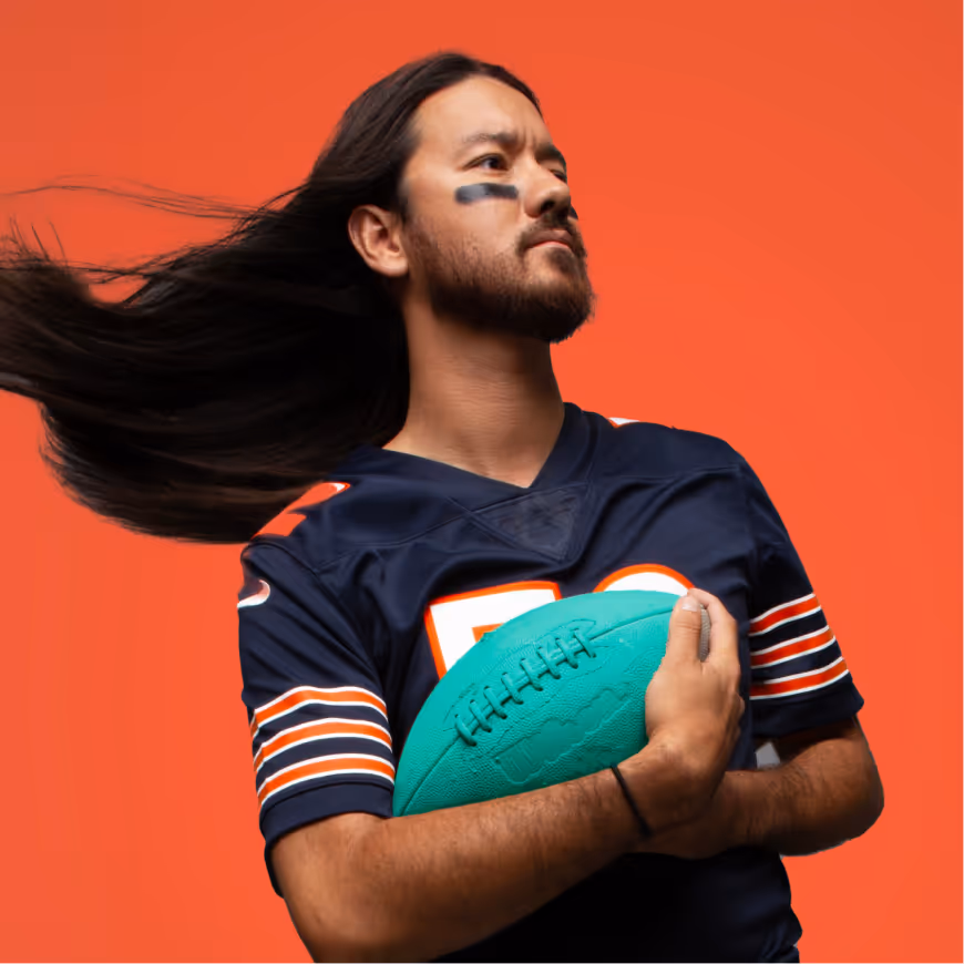 Man with long hair flowing, wearing a navy football jersey, holding a teal football against an orange background.
