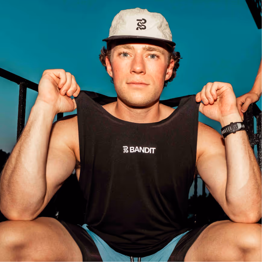 Young man wearing a white cap and black sleeveless shirt with 'BANDIT' logo, holding the shirt collar while sitting outdoors.