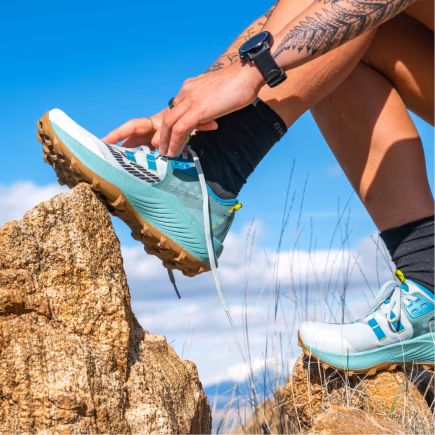 Person with tattooed arm and smartwatch putting on teal and white trail running shoes while sitting on rocks under a blue sky.