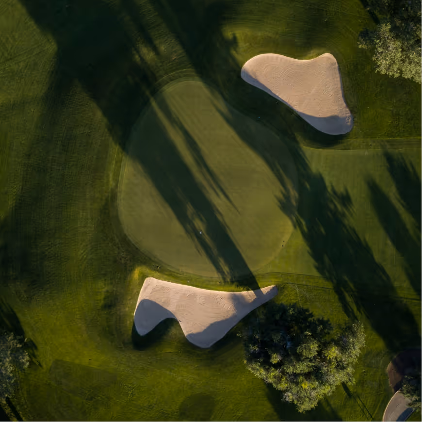 Aerial view of a golf green with two sand bunkers and shadows of tall trees on the grass.