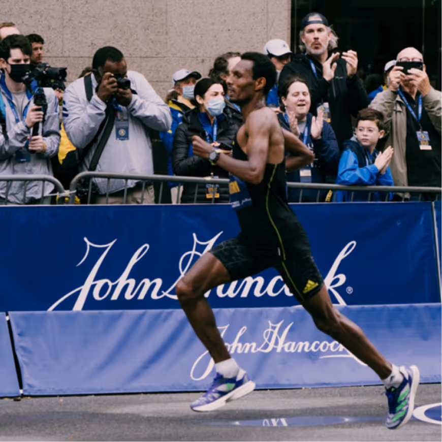Male runner in black athletic gear sprinting past a John Hancock branded barrier with spectators taking photos and clapping behind him.