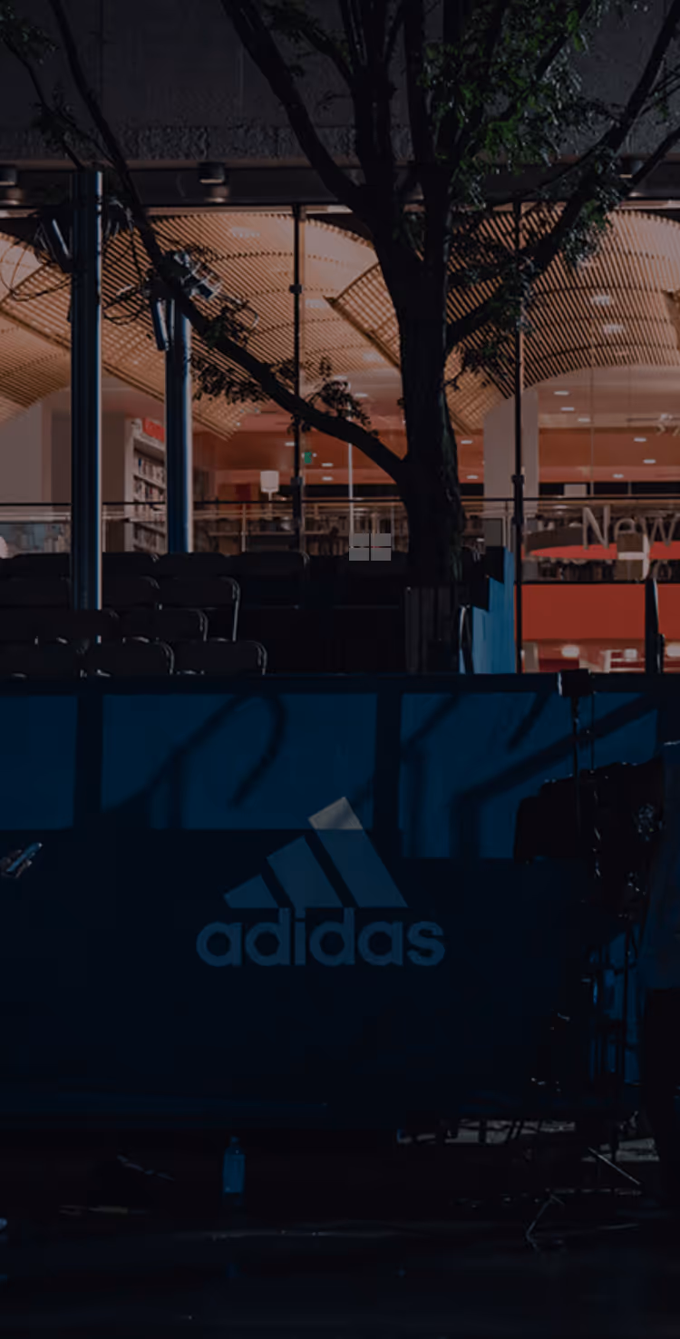 Dimly lit outdoor area at night with a large tree in front of a glass building interior and Adidas logo on a dark banner.