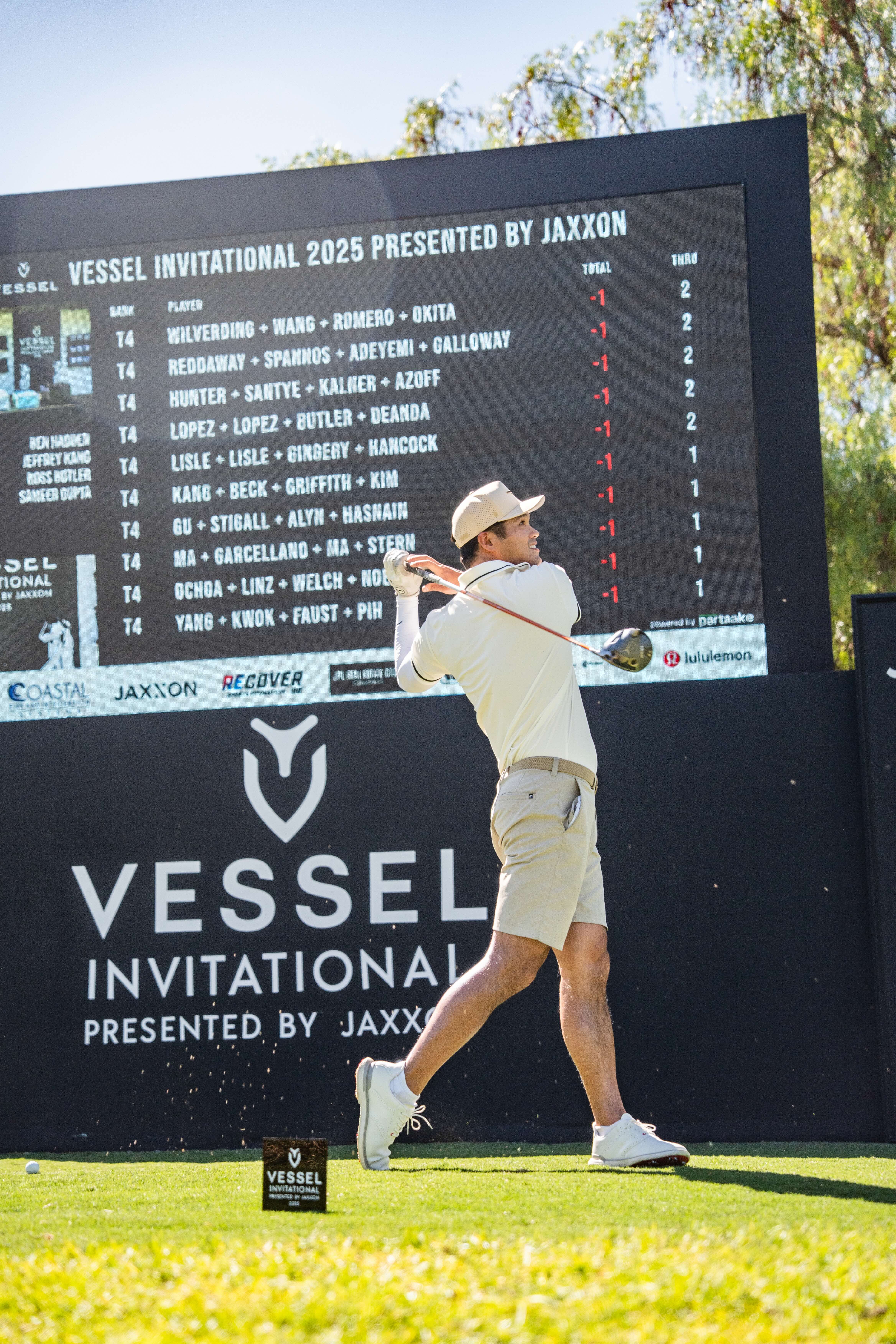 Smiling golfer holding a club on a sunny golf course with other players and golf carts in the background.