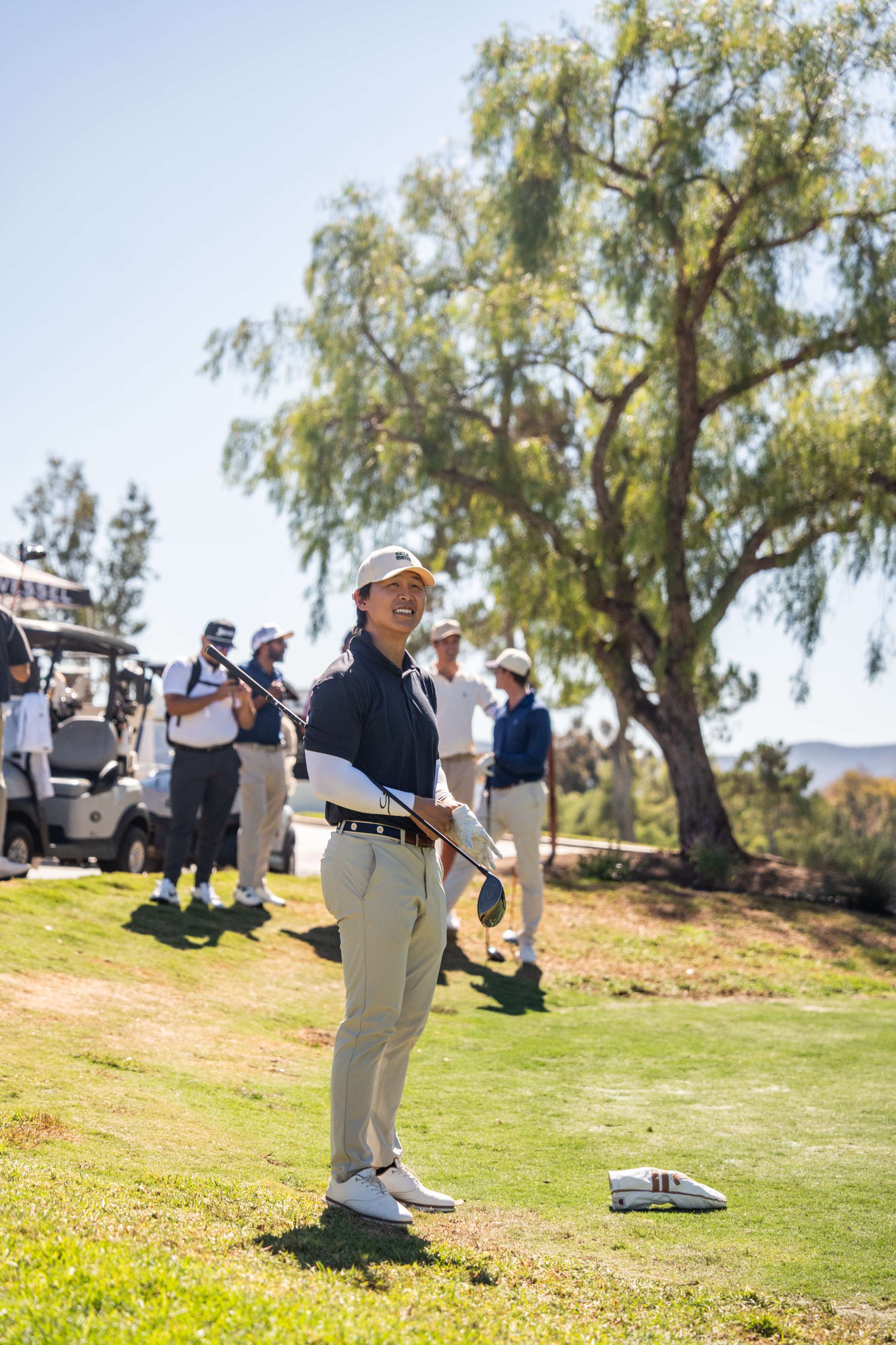 Smiling golfer holding a club on a sunny golf course with other players and golf carts in the background.