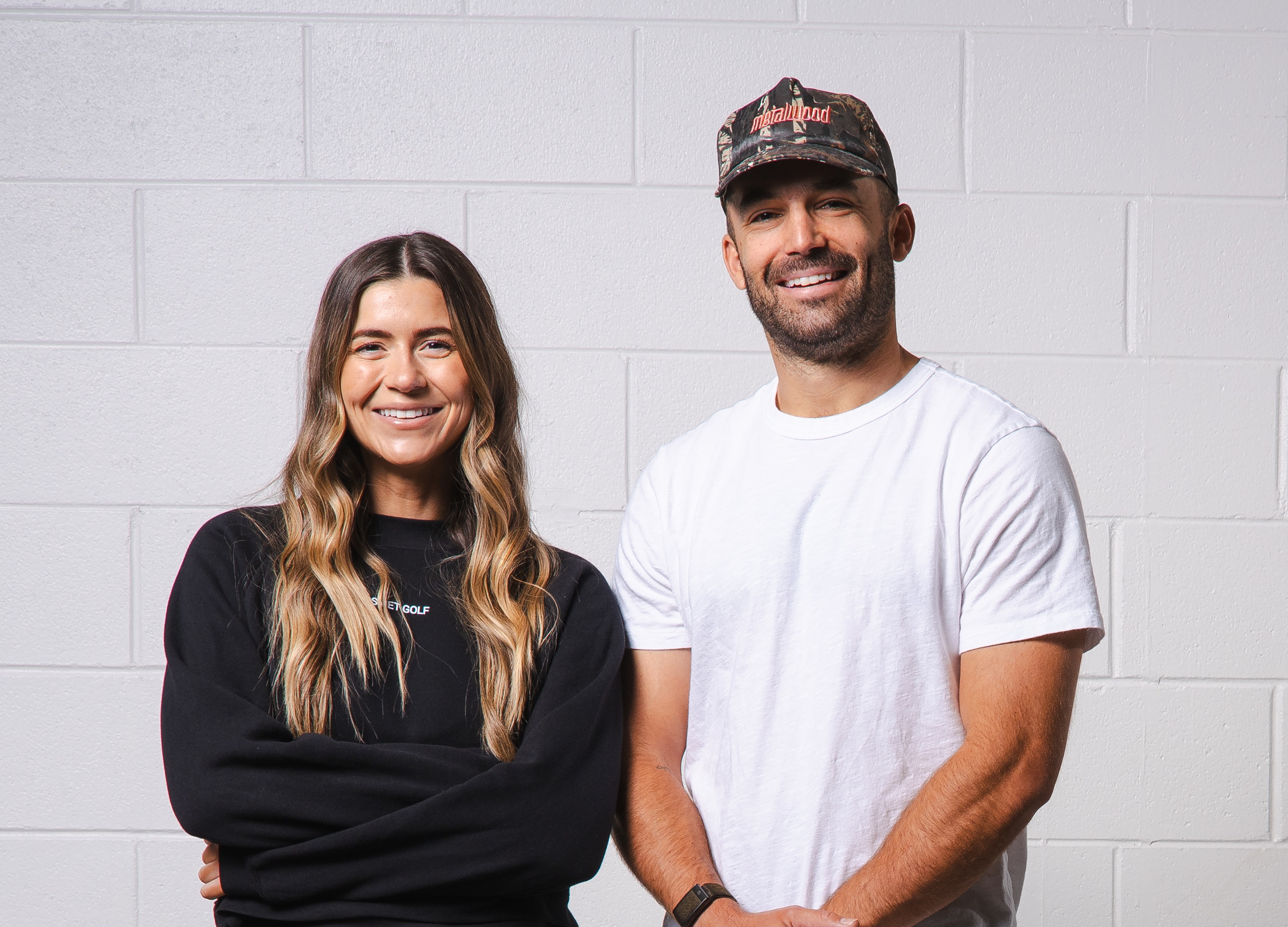 Smiling woman in black sweatshirt standing with arms crossed next to smiling man in white t-shirt and camouflage cap against a white brick wall.
