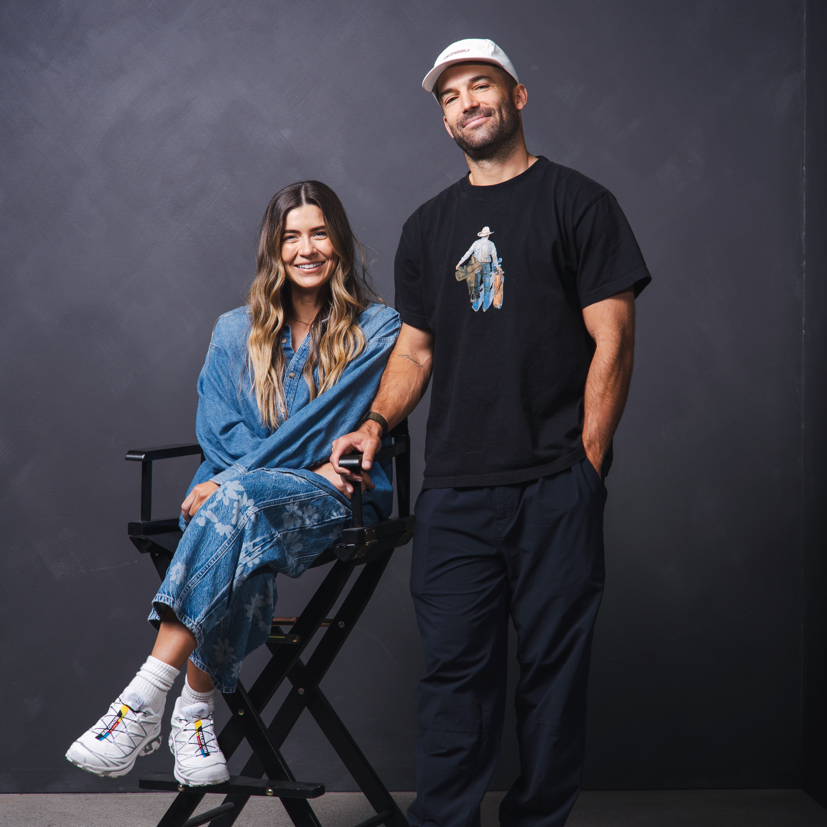 Smiling woman in denim outfit sitting on a director's chair next to a man wearing a black t-shirt and white cap against a dark gray backdrop.