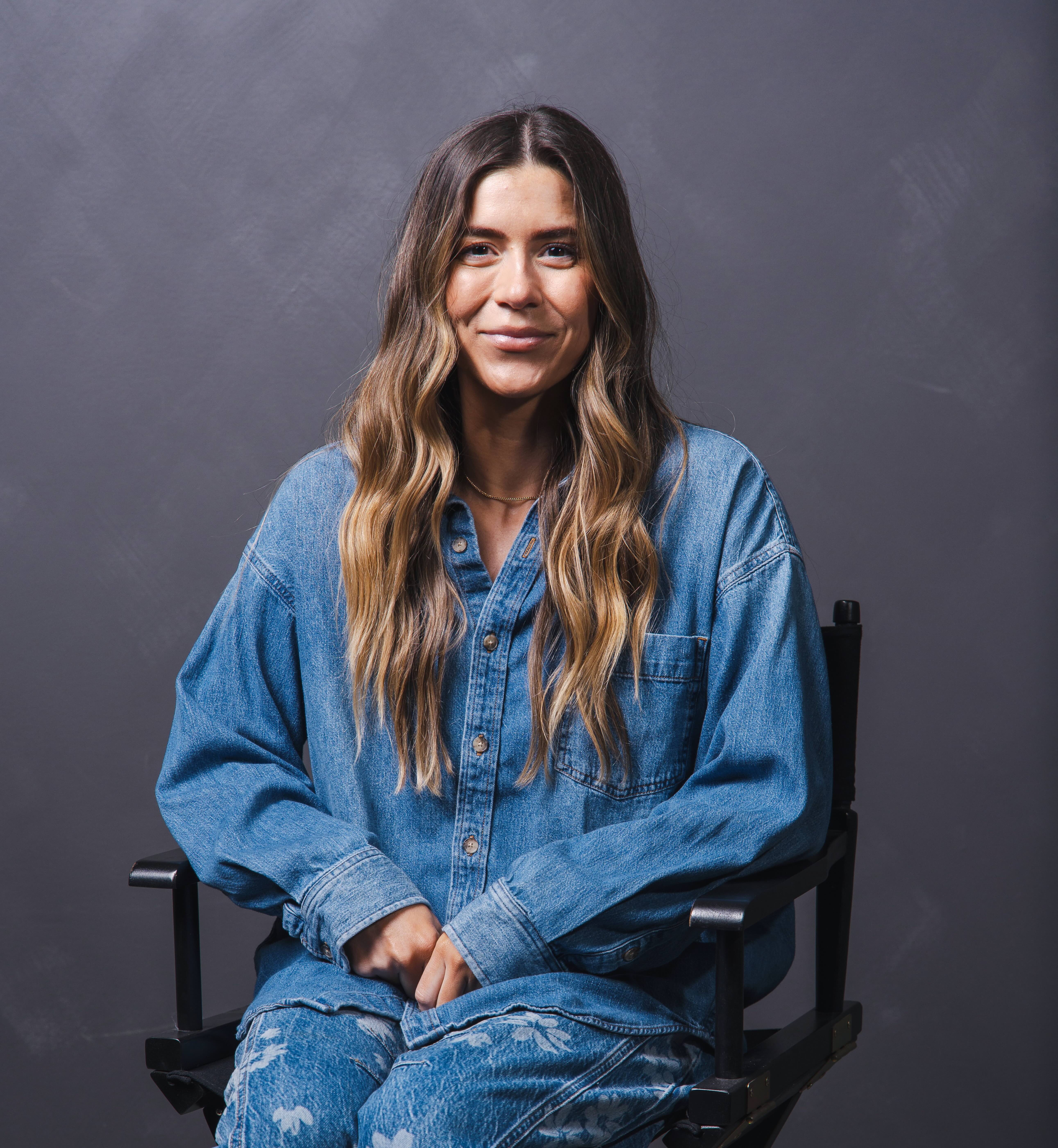 Woman with long wavy hair wearing a blue denim shirt and matching pants, sitting on a black chair against a gray background.