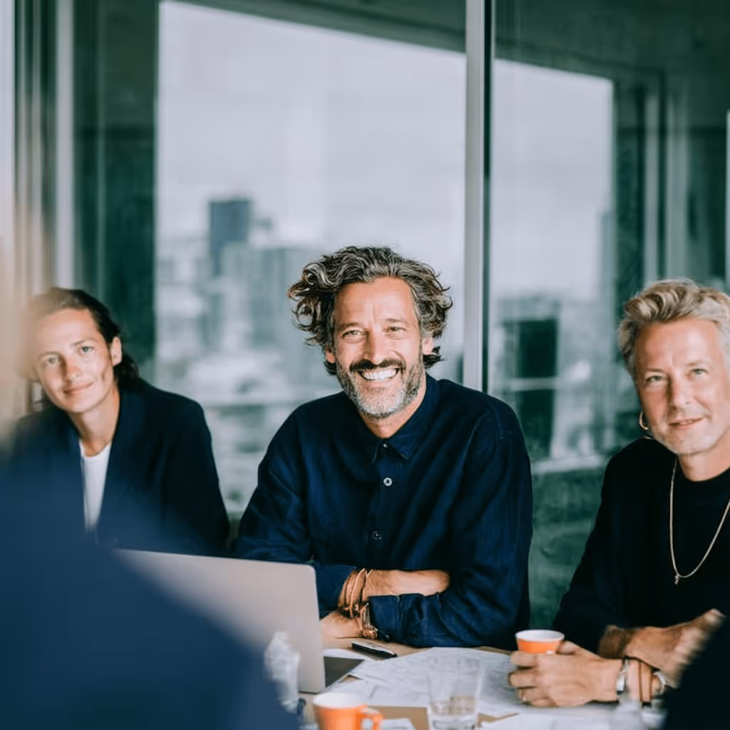Trois professionnels souriants assis à une table de réunion avec un ordinateur portable et des tasses orange.