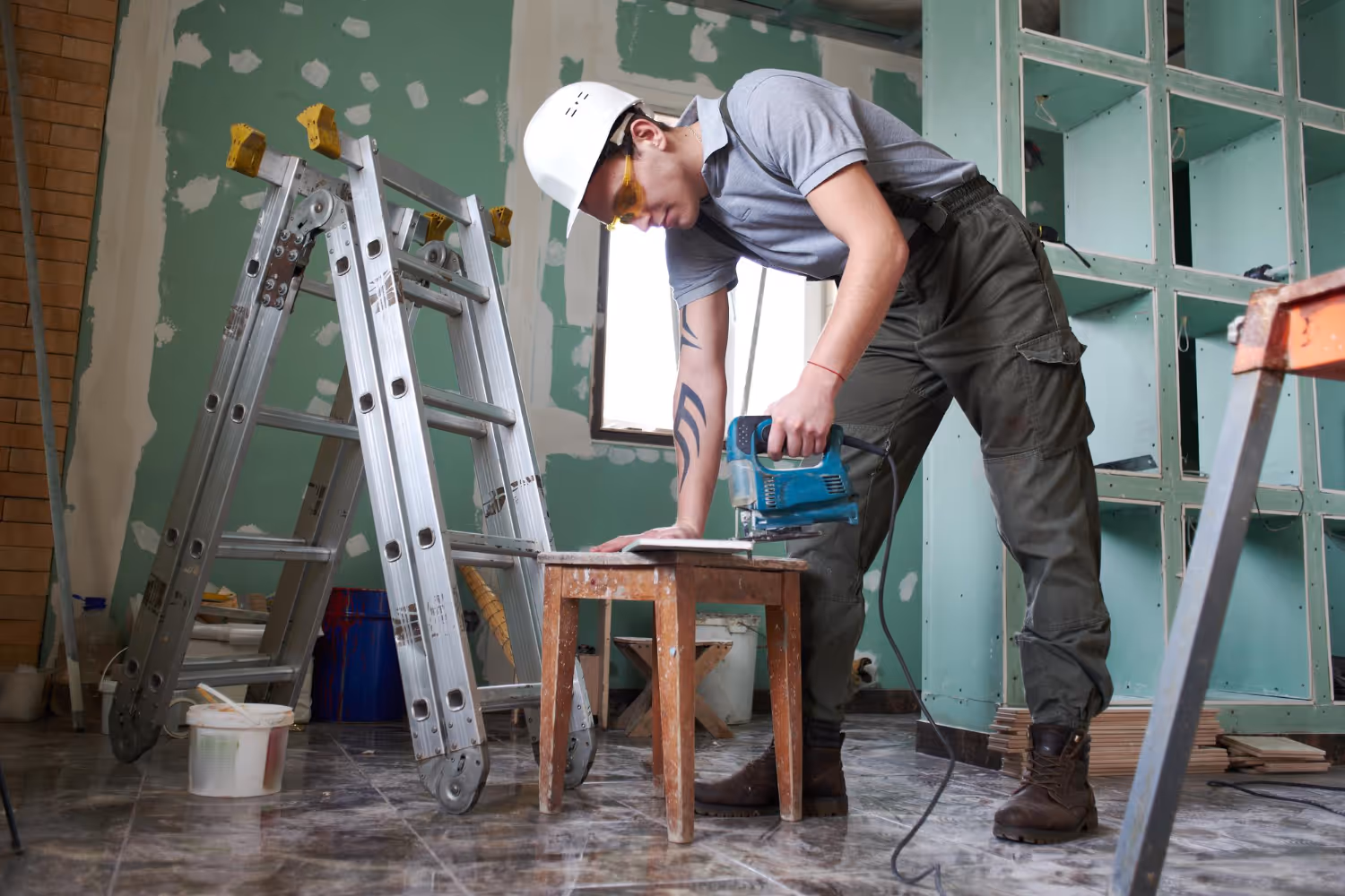 Construction worker in protective gear using a jigsaw to cut material on a small wooden table inside a room under renovation.