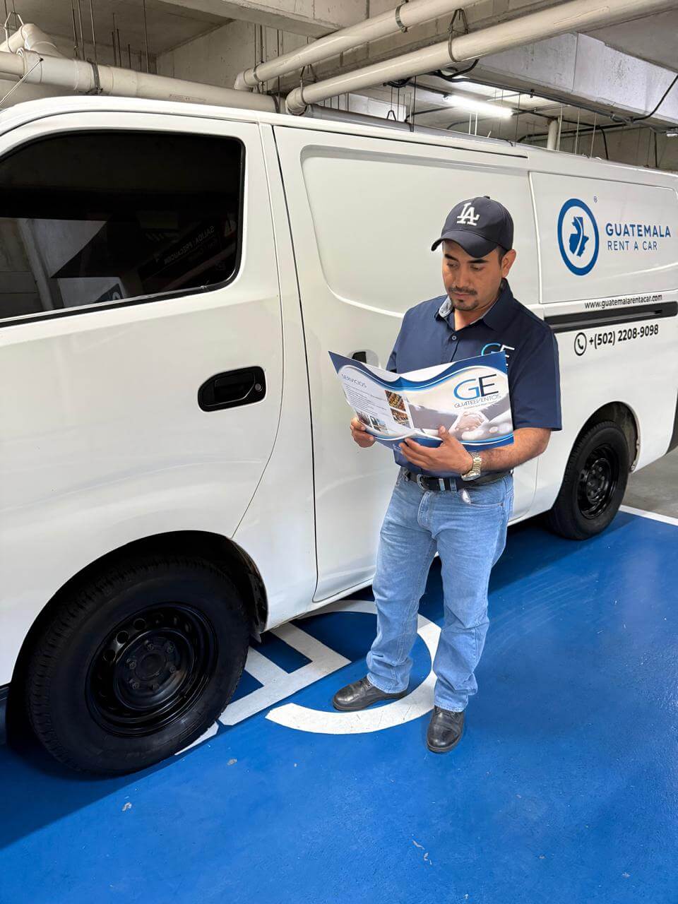 Man in navy shirt and cap reading a brochure while standing next to a white van with Guatemala Rent A Car logo in a parking garage.
