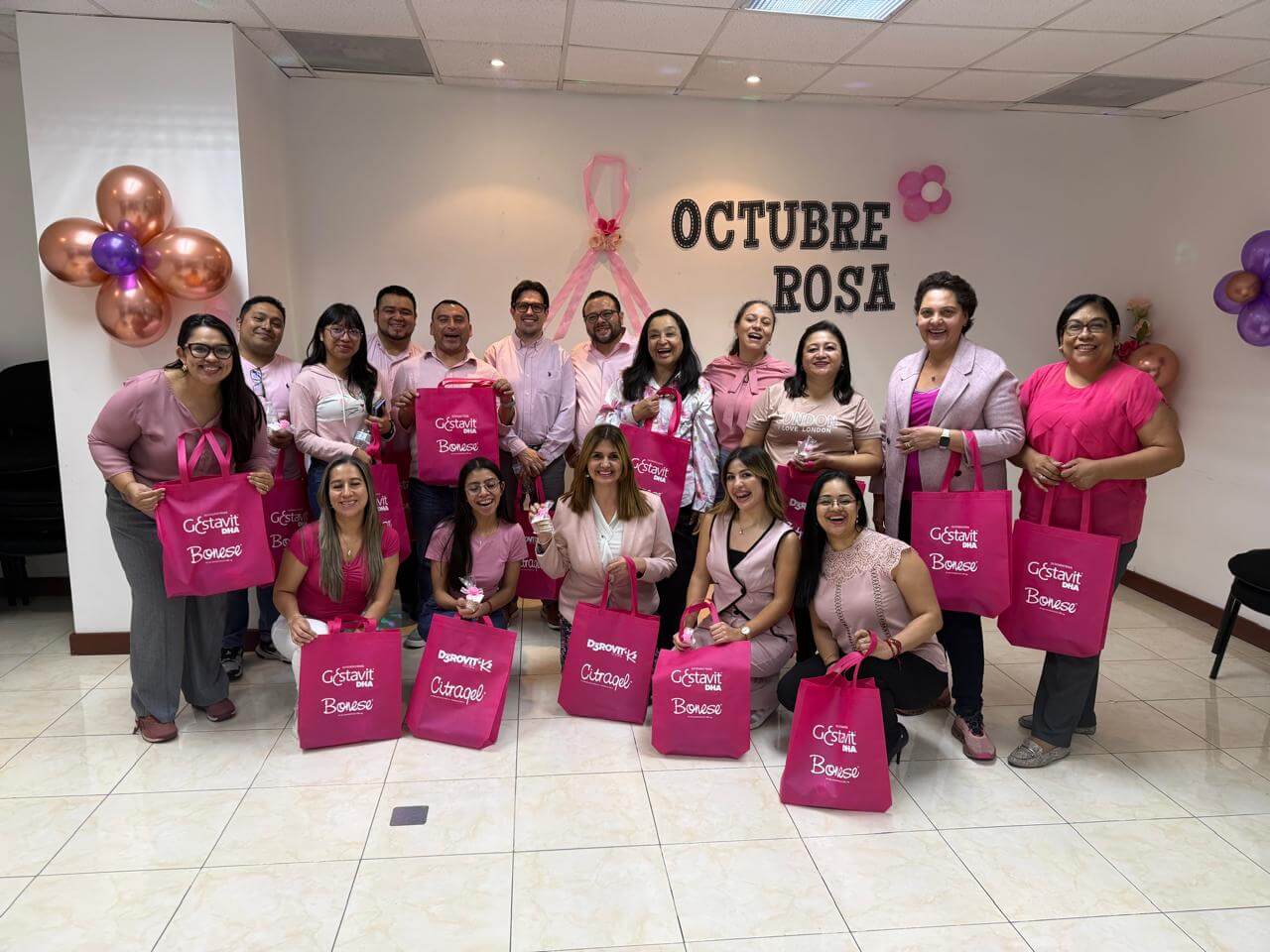 Group of women and men dressed in pink holding pink gift bags and standing in front of a wall decorated with a pink ribbon and letters spelling 'OCTUBRE ROSA'.