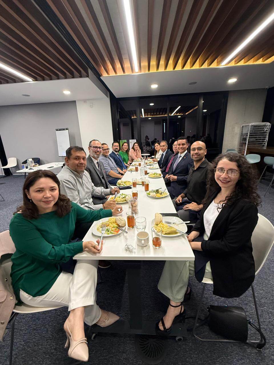 A group of twelve people sitting around a long table in an office setting, smiling and having a meal together.