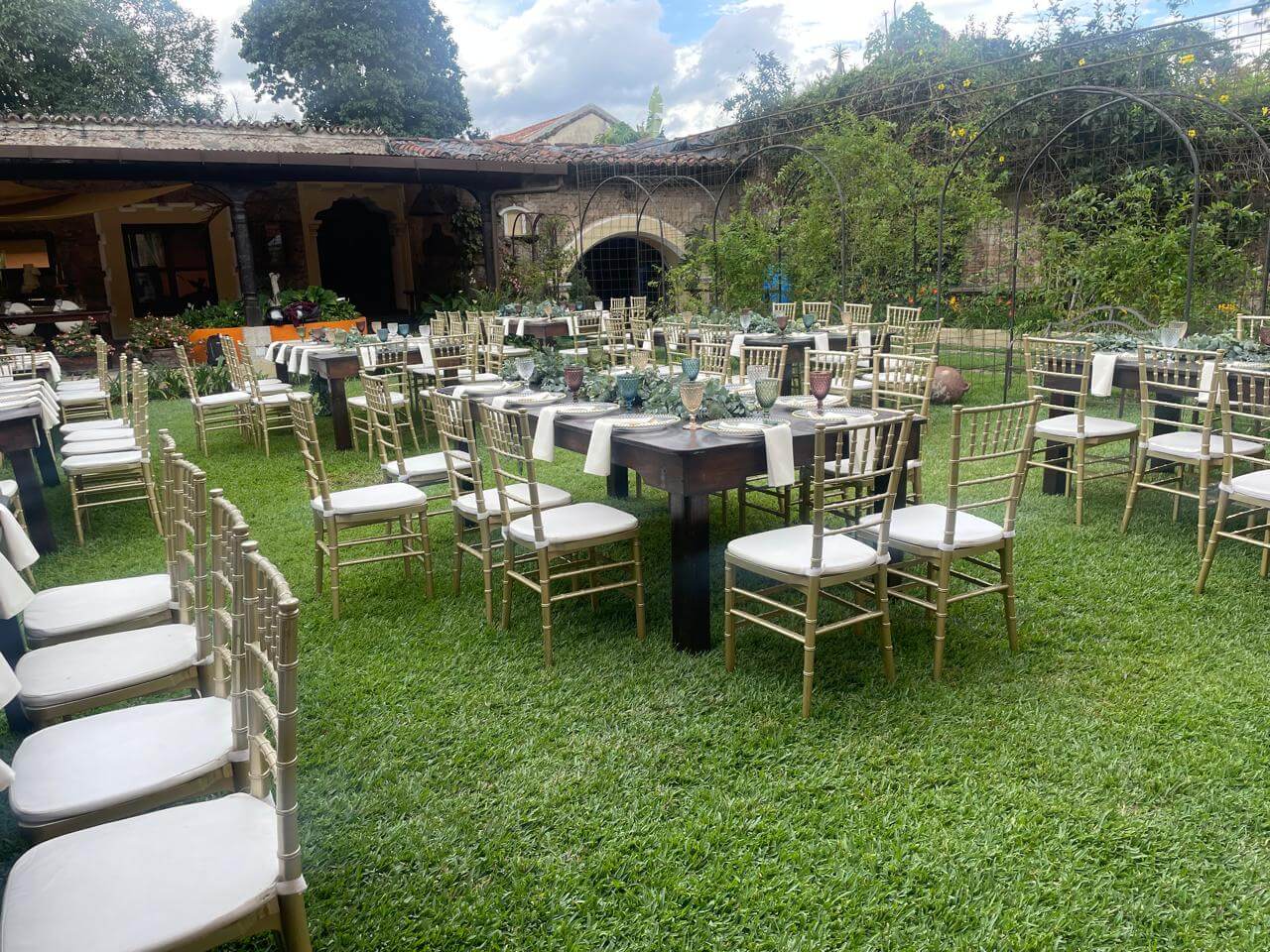 Outdoor garden setup with multiple wooden tables and beige cushioned chairs arranged on green grass.