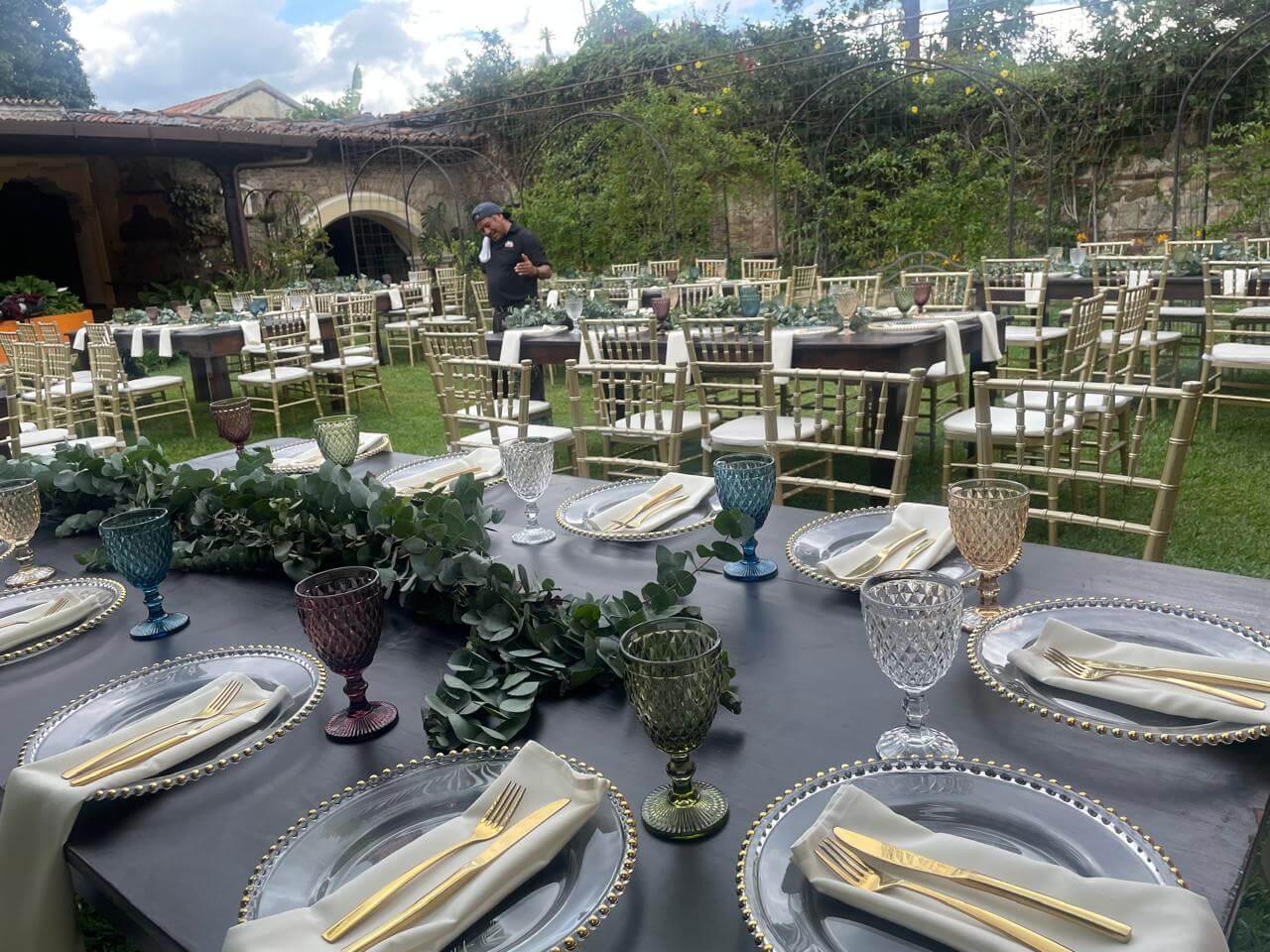 Outdoor event setup with multiple tables dressed in gray tablecloths, clear plates with gold-rimmed edges, gold cutlery on white napkins, and colorful glass goblets, with gold chairs on green grass and greenery in the background.