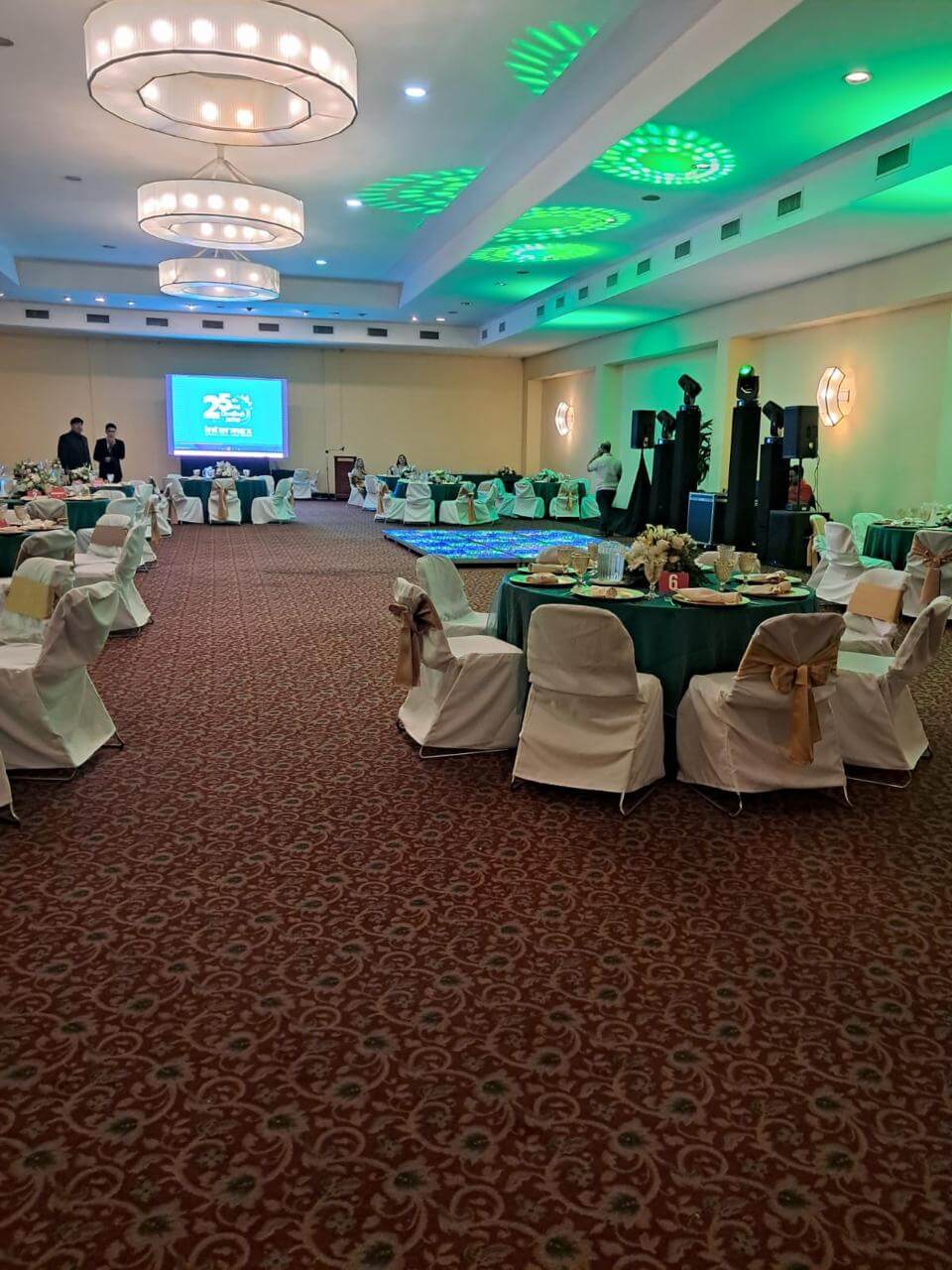 Indoor banquet hall with round tables covered in green tablecloths, white chairs with gold bows, ceiling chandeliers, and a screen displaying a presentation.