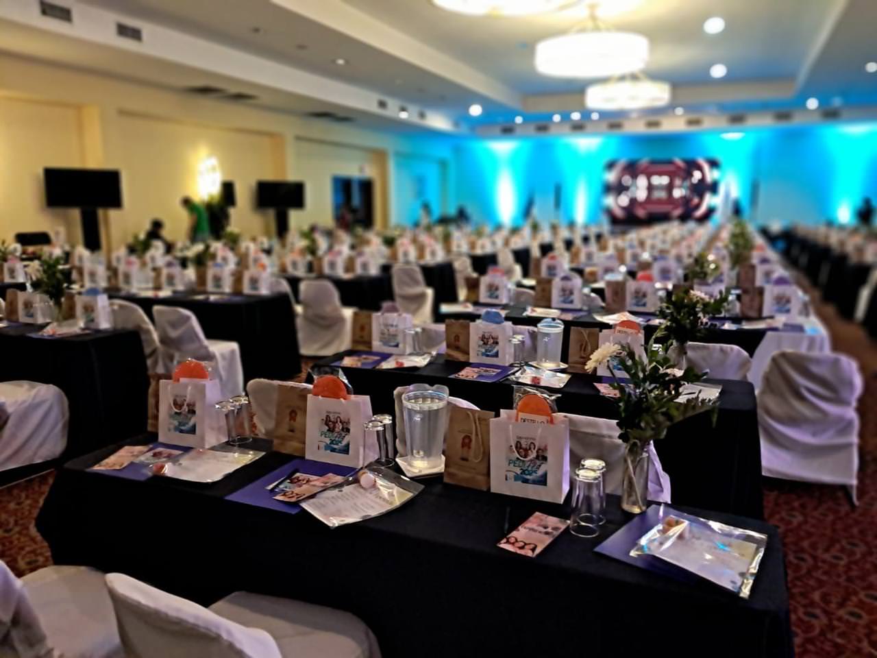 Large conference room with rows of tables and chairs covered in white cloth, each table set with gift bags, flowers, and glassware.