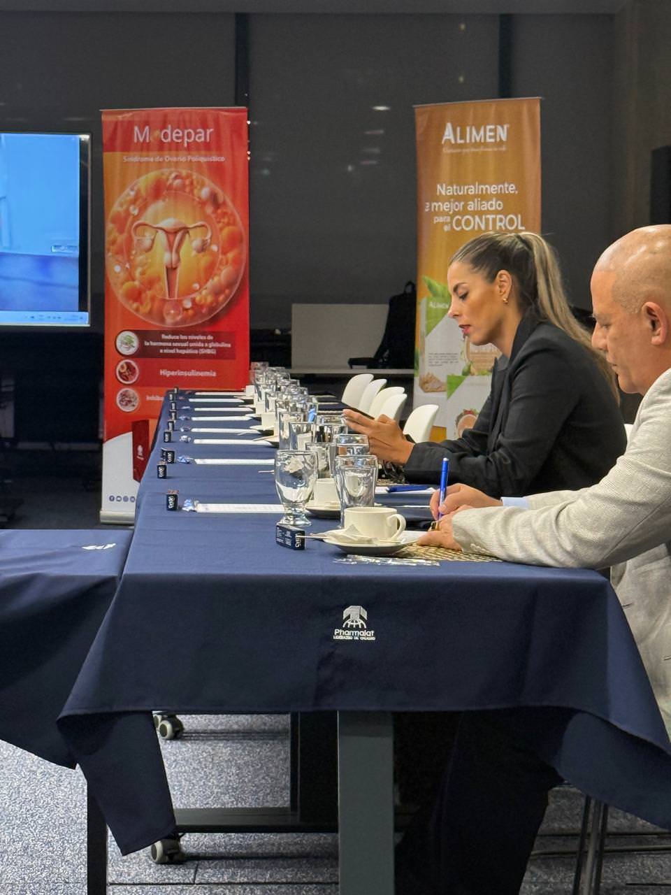 Two people seated at a long table with blue cloth in a conference room, one writing with a pen and the other looking at a phone, with promotional banners in the background.