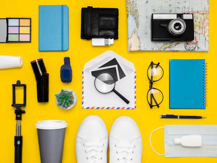Flat lay of travel and personal items on yellow background including white sneakers, camera on map, glasses, notebook, magnifying glass over envelope, coffee cup, and face mask with sanitizer.