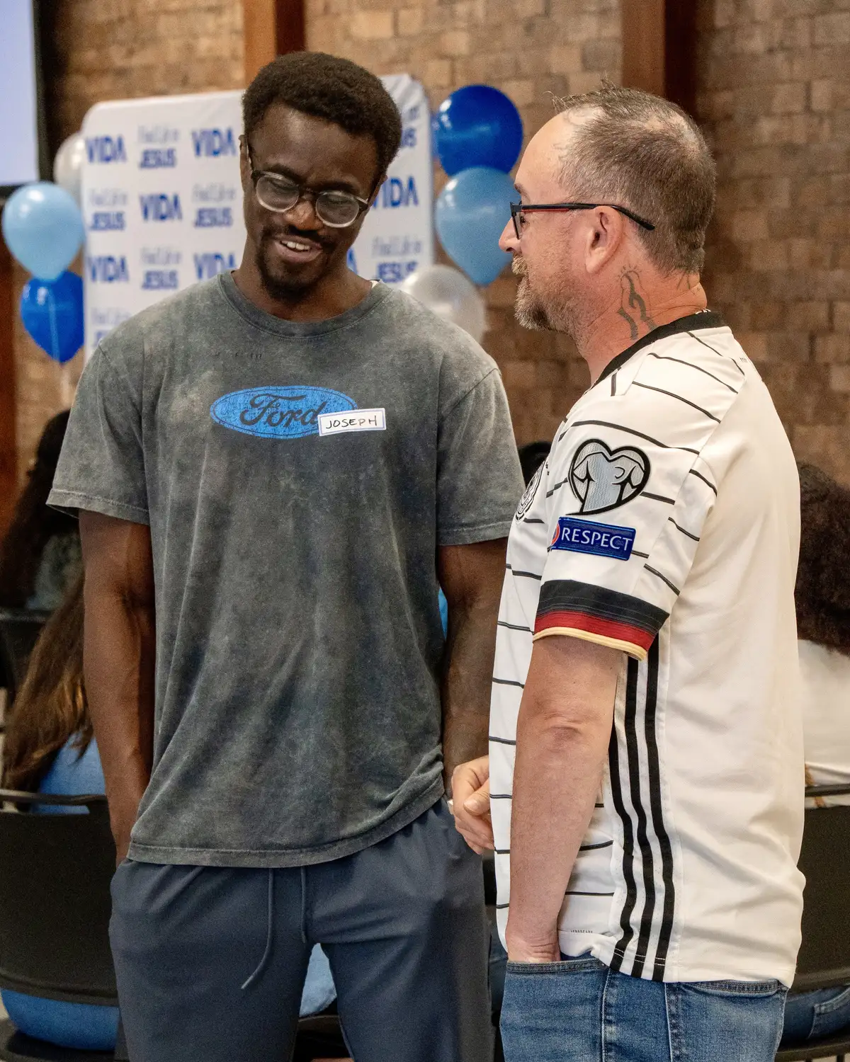 Two men smiling and talking indoors near blue and white balloons and a Vida Jesus poster.