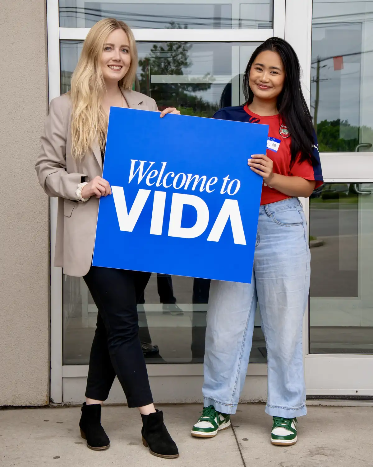 Two women smiling and holding a blue sign that says 'Welcome to VIDA' in front of a glass door.
