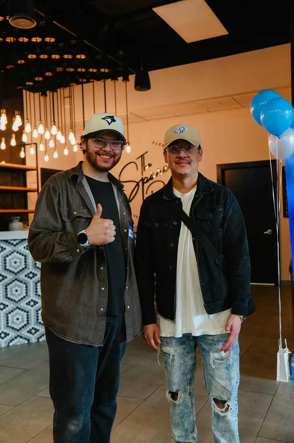 Two men standing indoors, one giving thumbs up, both wearing caps and casual outfits with glasses.