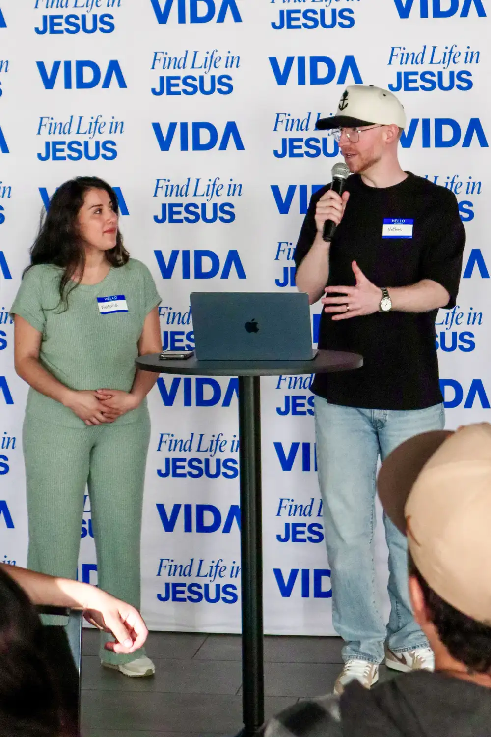 Man in a cap speaking into a microphone beside a woman standing near a laptop and table at event.