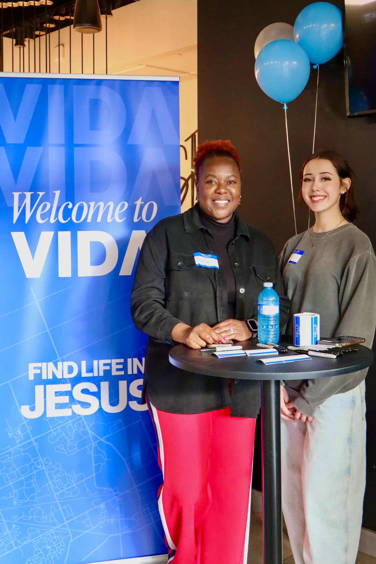 Two smiling women standing behind a tall round table with pens, name tags, a water bottle, and a roll of "Hello" stickers. Behind them is a blue sign that reads "Welcome to VIDA. Find Life in Jesus." Three balloons, two blue and one white, float beside the women.