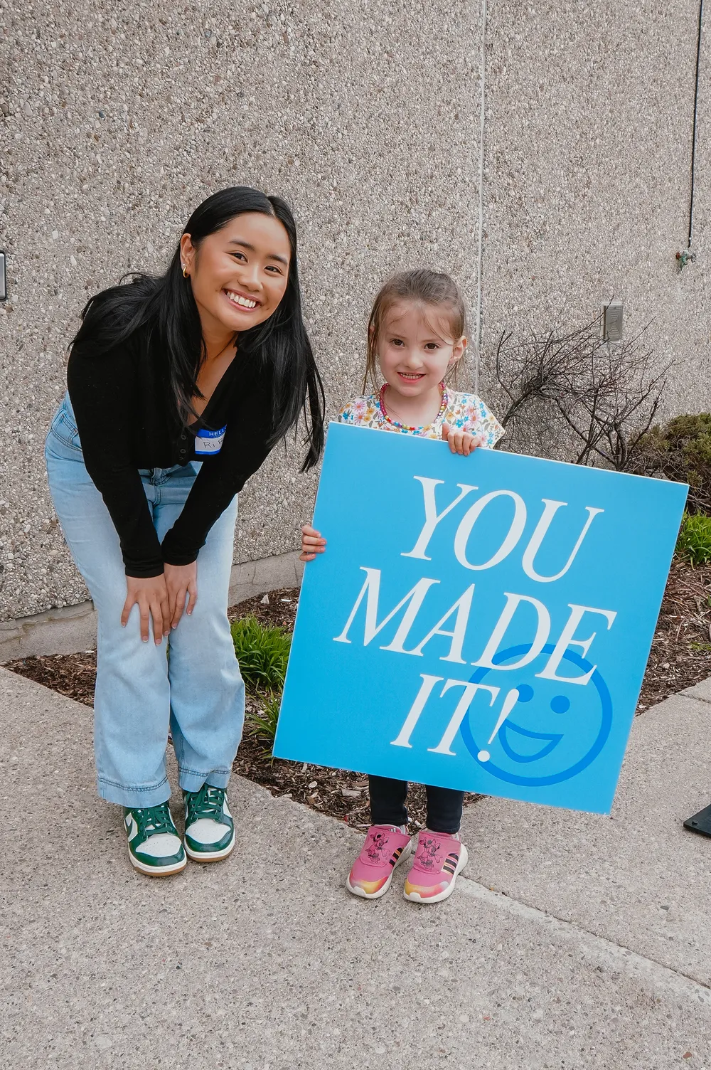 Smiling young woman with long black hair wearing a black top, blue jeans, and green and white sneakers stands next to a young girl holding a large blue sign that says 'YOU MADE IT!' with a smiley face on a sidewalk in front of a textured concrete wall.