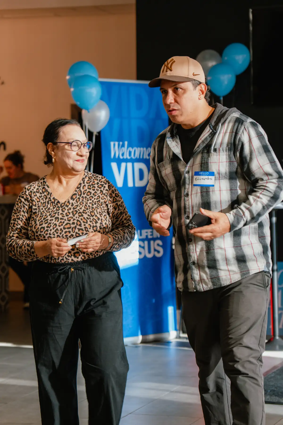 A man and a woman engaged in conversation indoors; the woman wears glasses, a leopard print top and black pants, the man wears a baseball cap, plaid shirt, and dark pants with a name tag that says Eduardo; blue and white balloons and a blue banner with white text are in the background.