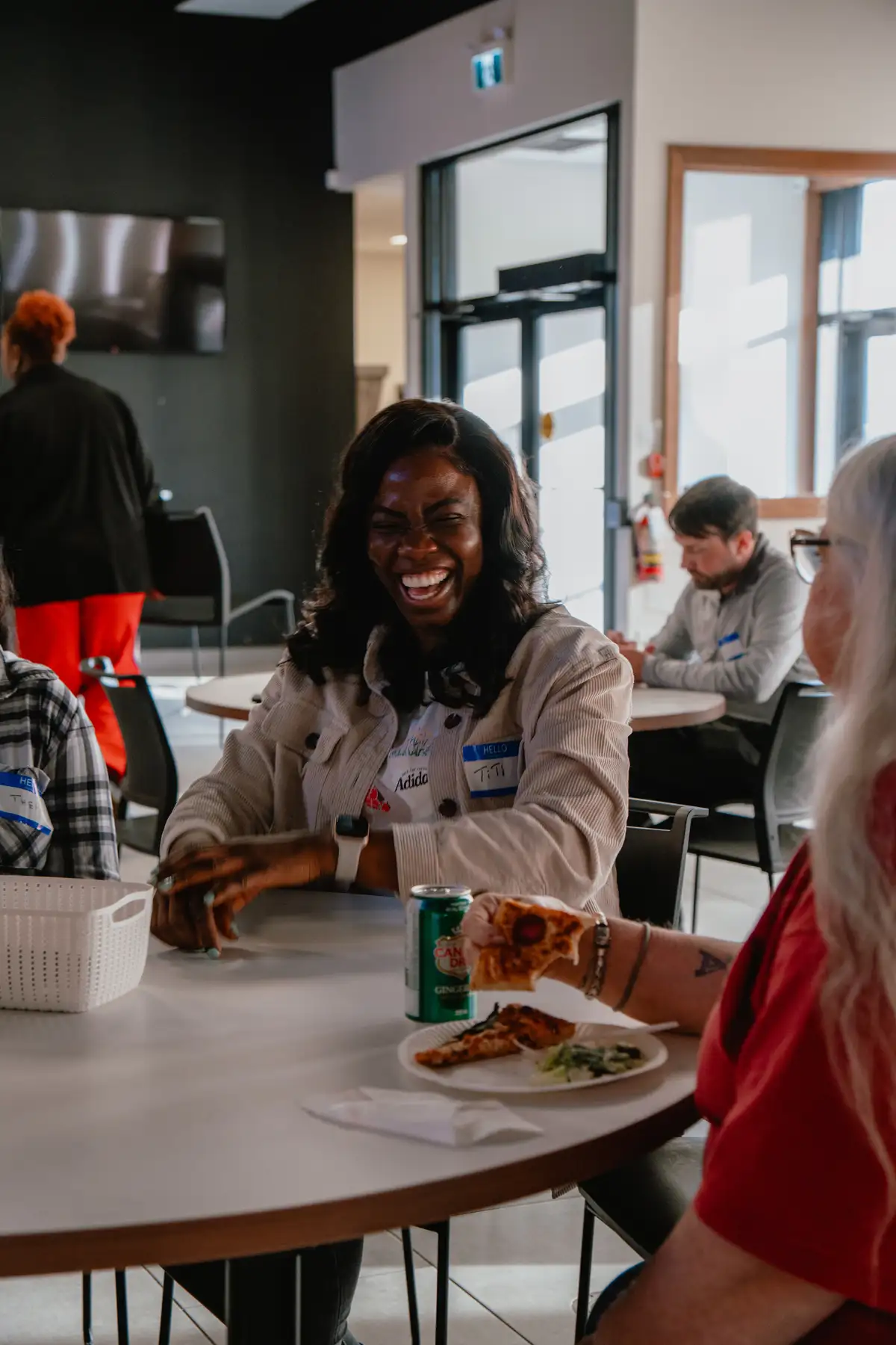 Smiling woman with a name tag sitting at a round table indoors with a can of ginger ale, a plate with pizza, and another person holding a slice of pizza.