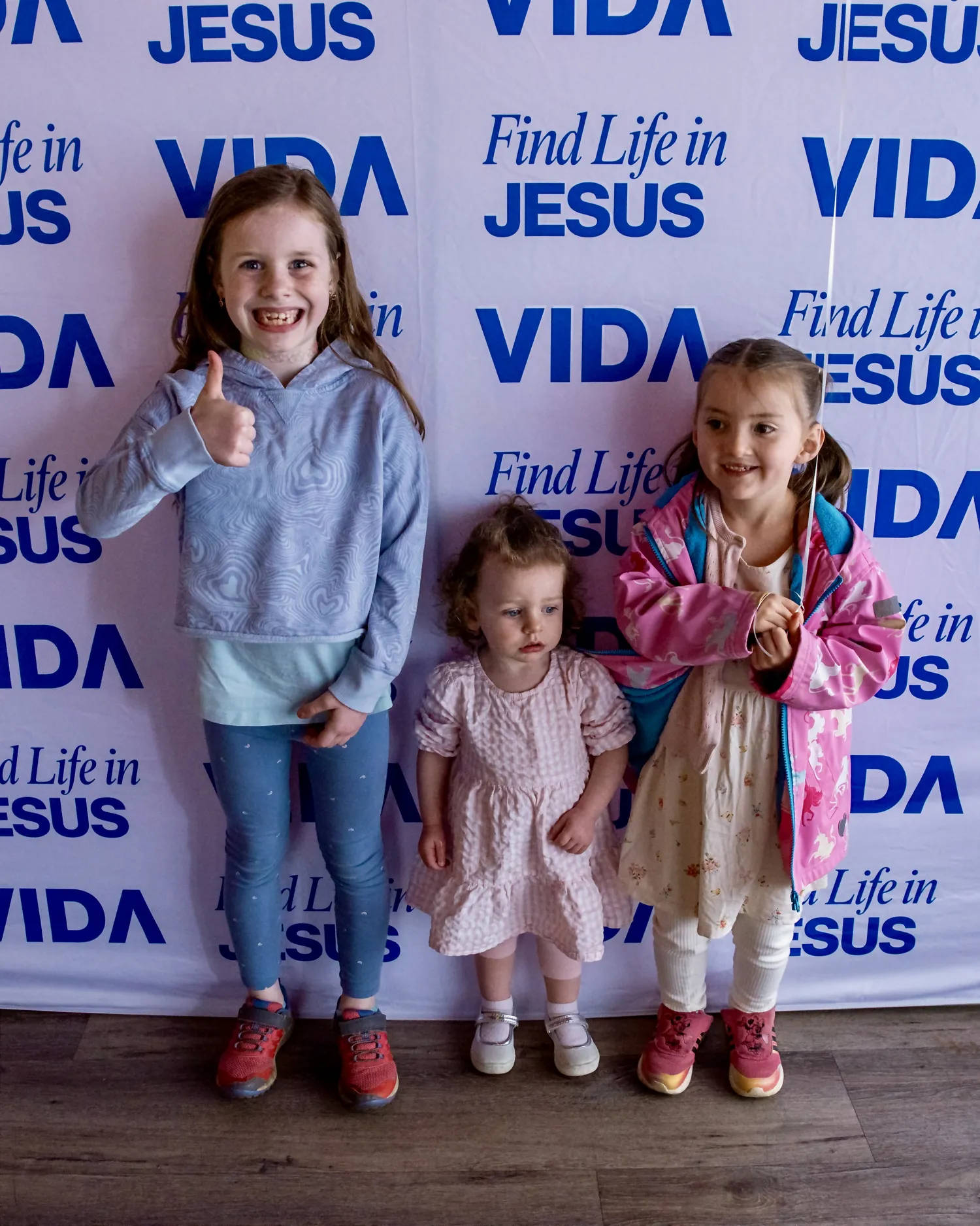 Three young girls standing indoors against a backdrop with repeated blue text reading "Find Life in JESUS" and "VIDA." The girl on the left wears a light blue hoodie, blue leggings, and red shoes, smiling with her thumb up. The middle girl wears a pale pink dress and white shoes, looking down. The girl on the right wears a pink jacket over a white dress, white tights, and pink shoes, holding a balloon string and smiling.