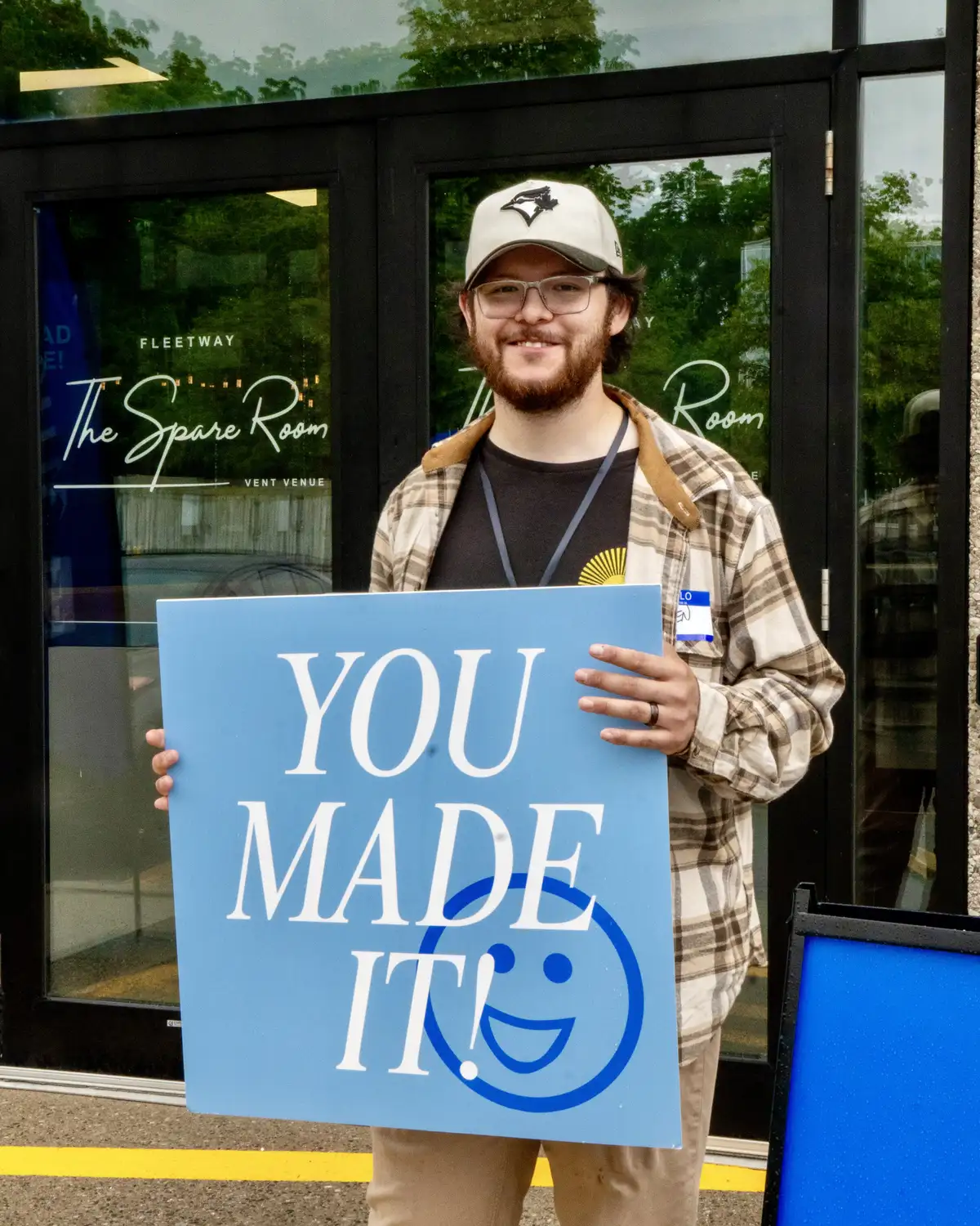 Man with glasses and a beard wearing a plaid shirt and baseball cap stands outside a venue holding a blue sign that reads 'YOU MADE IT!' with a smiley face.