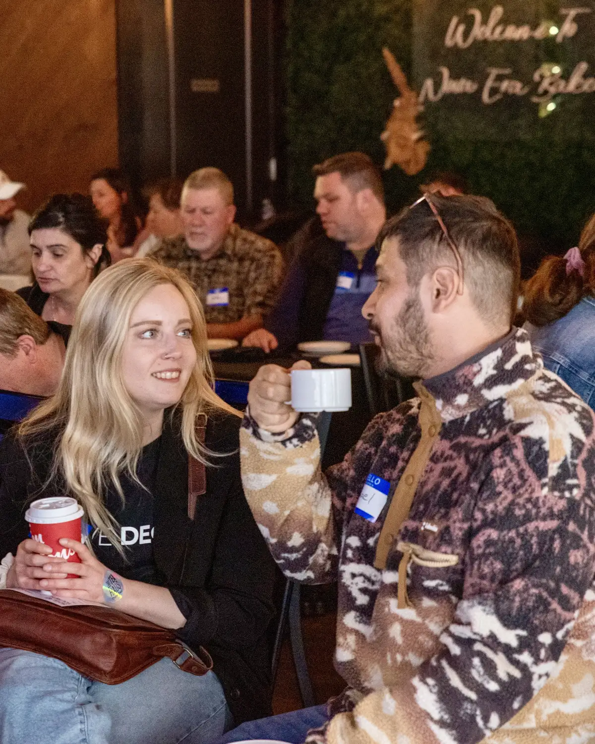 A man and a woman sitting and chatting in a crowded indoor setting, each holding a cup, with other people seated and engaged in the background, some wearing name tags.