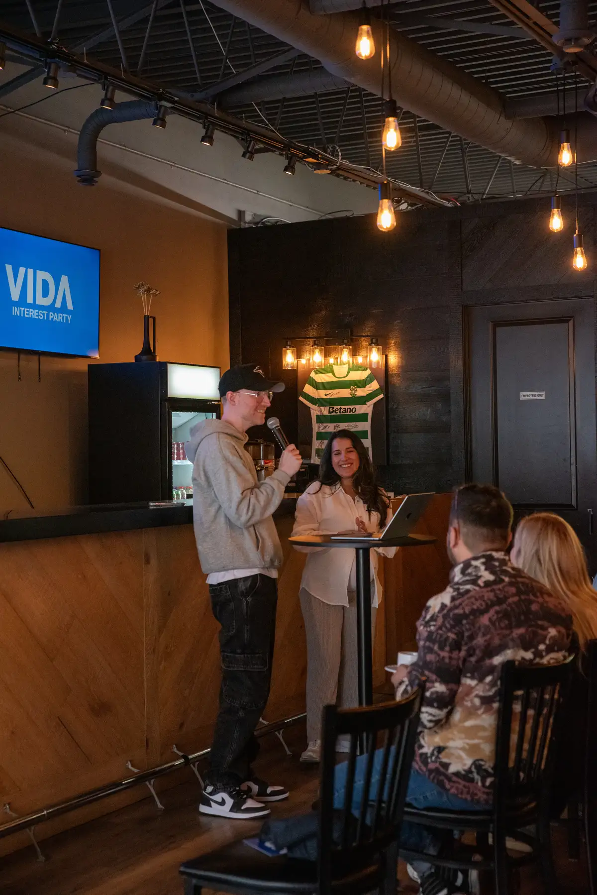 A man in a gray hoodie and black cap speaking into a microphone next to a woman in a white blouse standing at a tall table with a laptop, while two seated people watch in a warmly lit room with a sign that reads 'VIDA INTEREST PARTY' on a screen.
