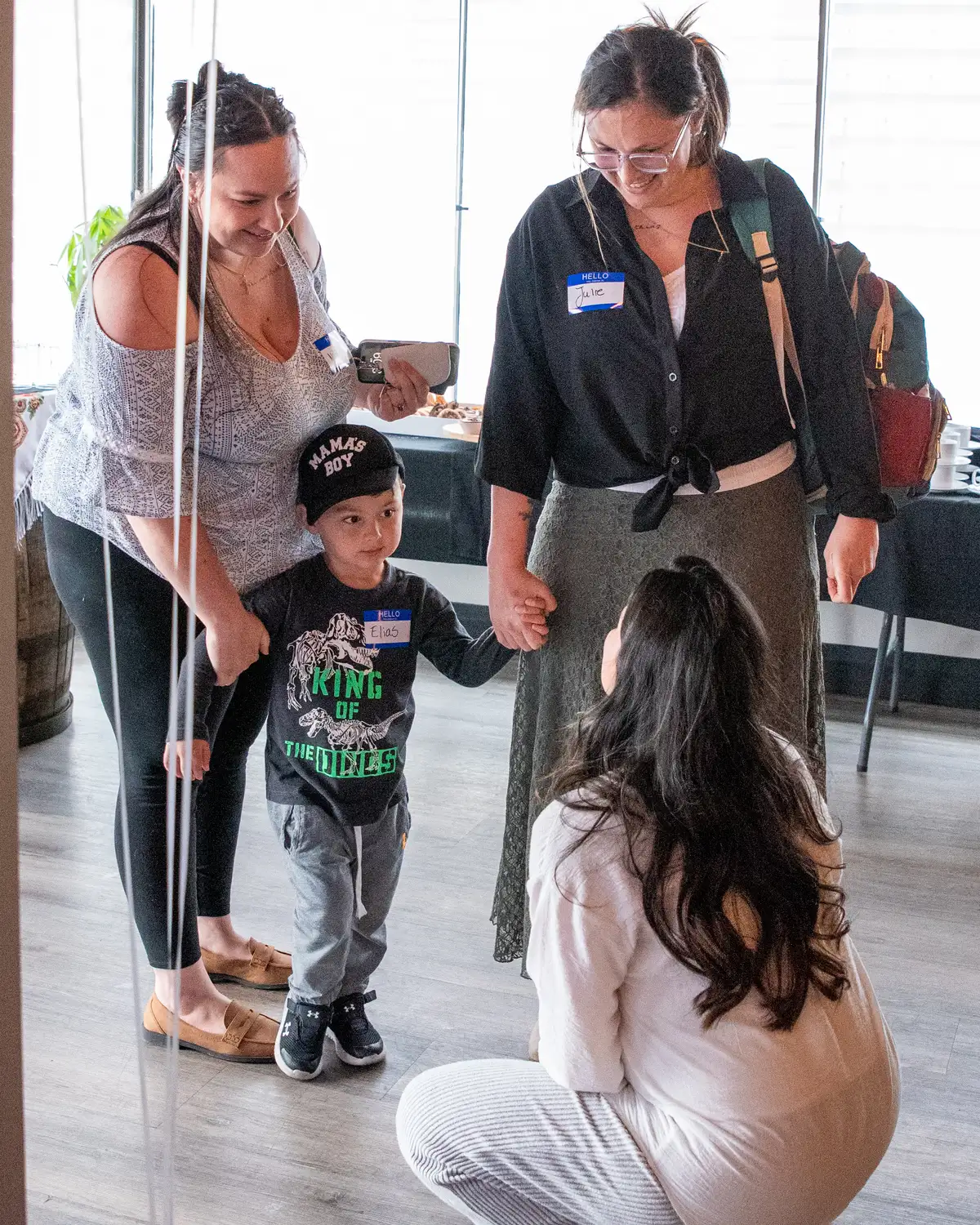 A young boy wearing a 'Mama's Boy' cap and 'King of the Dinos' shirt holds hands with two women, one standing on each side, while a third woman crouches in front of them, all smiling and interacting in a bright room with wooden floors.