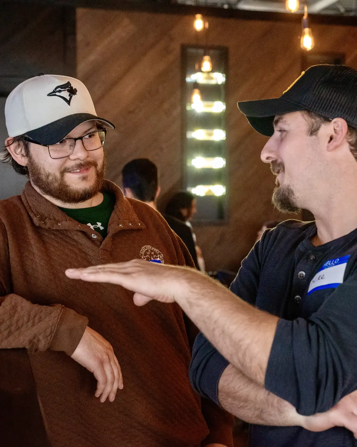 Two men having a conversation indoors; one wearing glasses, a white and black baseball cap, and a brown sweater with a patch, the other with a black baseball cap and dark shirt gestures with his hand.