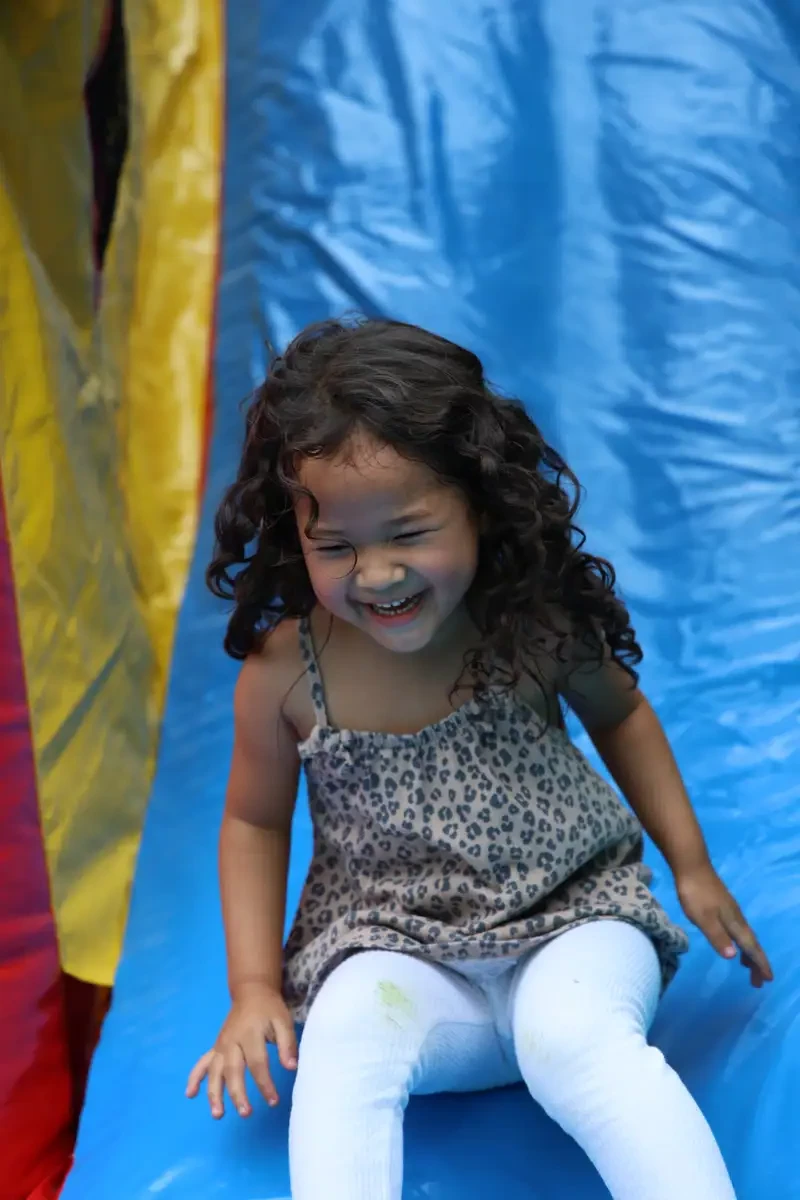 Smiling young girl with curly hair wearing a leopard print dress and white leggings sliding down a blue inflatable slide.