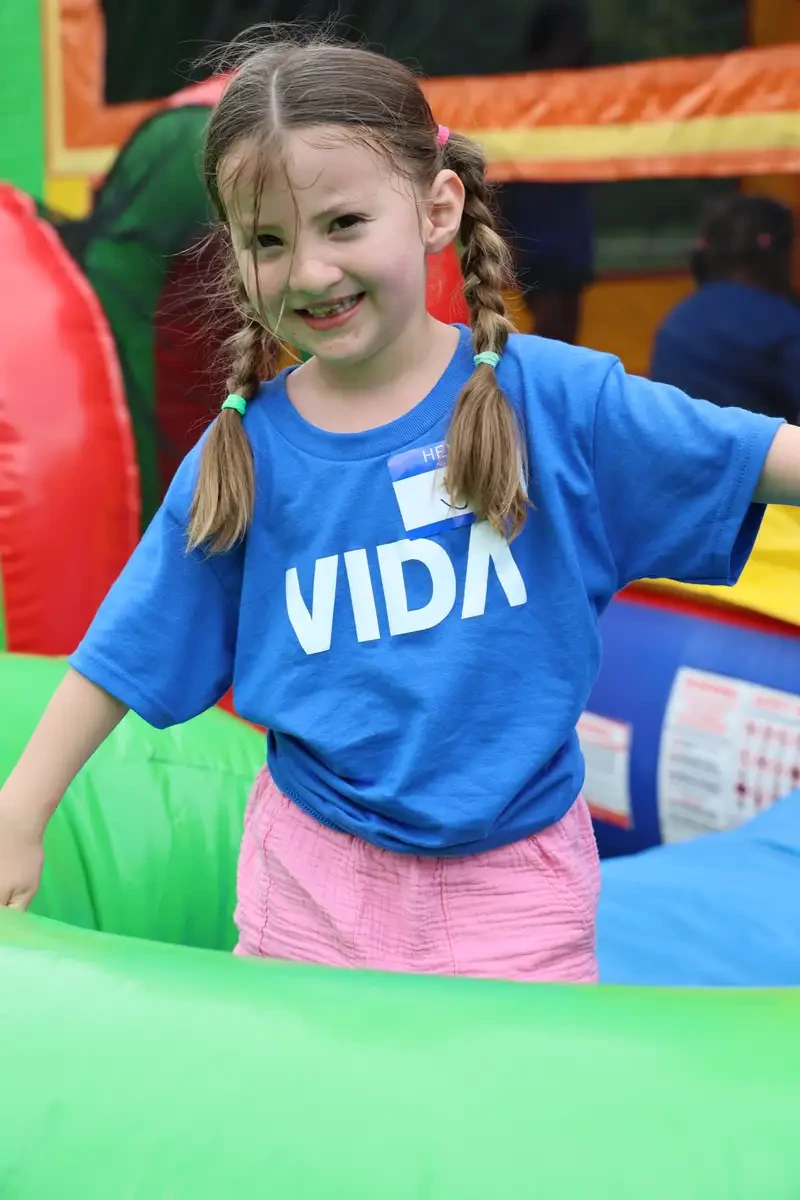 Smiling young girl with braided hair wearing a blue shirt with 'VIDA' printed on it and pink shorts, playing on a colorful inflatable bounce house.