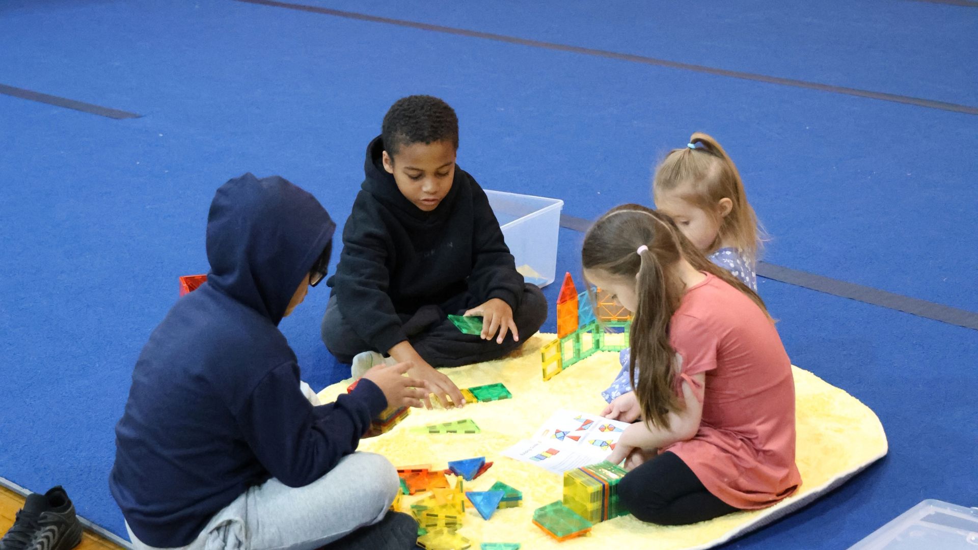Four children sitting on a yellow mat on a blue floor, playing and building with colorful translucent geometric shapes, one child holding an instruction sheet.