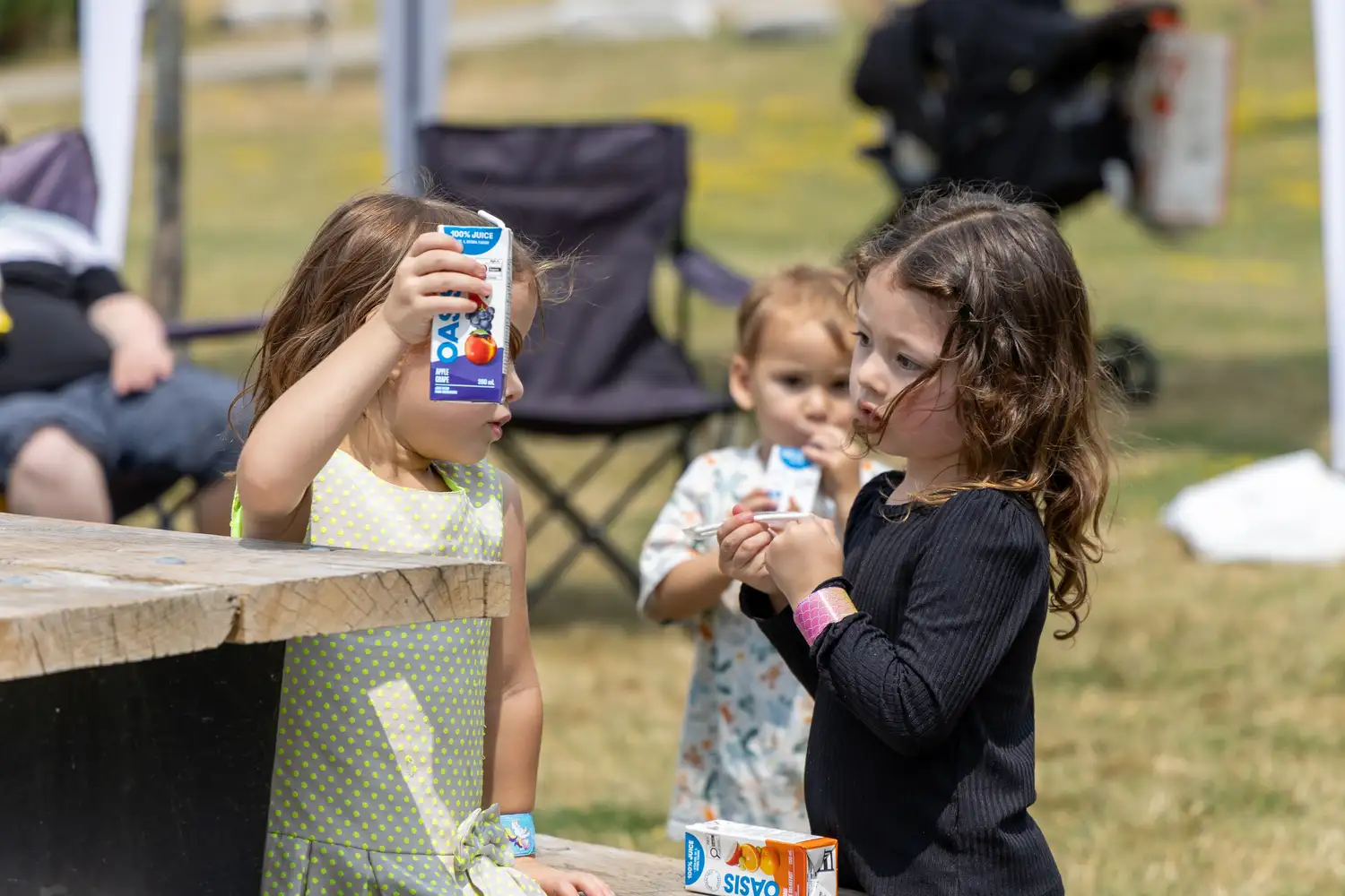 Three young children outdoors on a sunny day, two girls in the foreground with one holding up a juice box and the other holding a straw, and a toddler in the background drinking from a juice box.