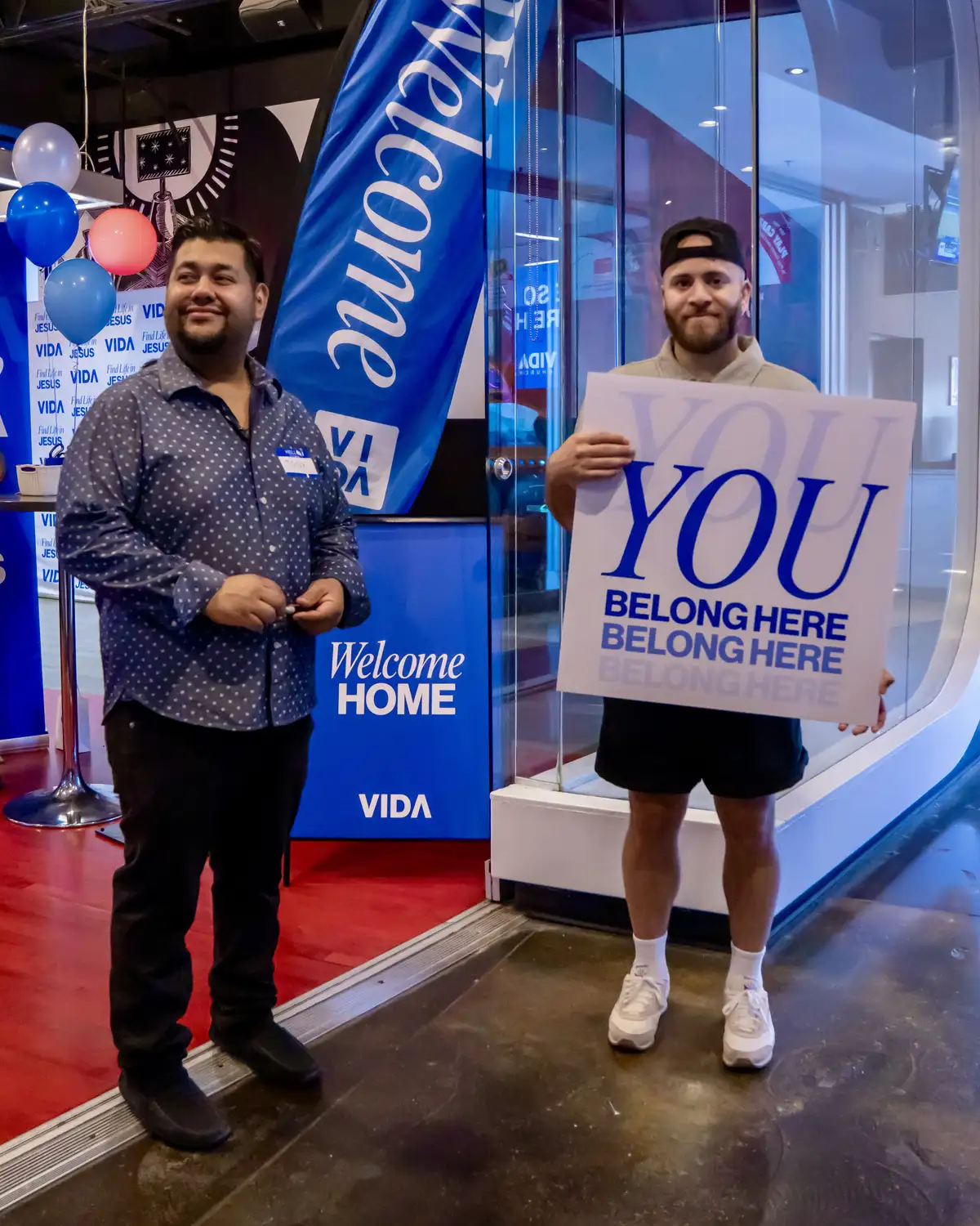 Two men standing indoors near a blue banner that says 'Welcome Home VIDA.' One man in a blue patterned shirt smiles while the other in a beige shirt, black shorts, white socks, and white sneakers holds a sign reading 'YOU BELONG HERE.'
