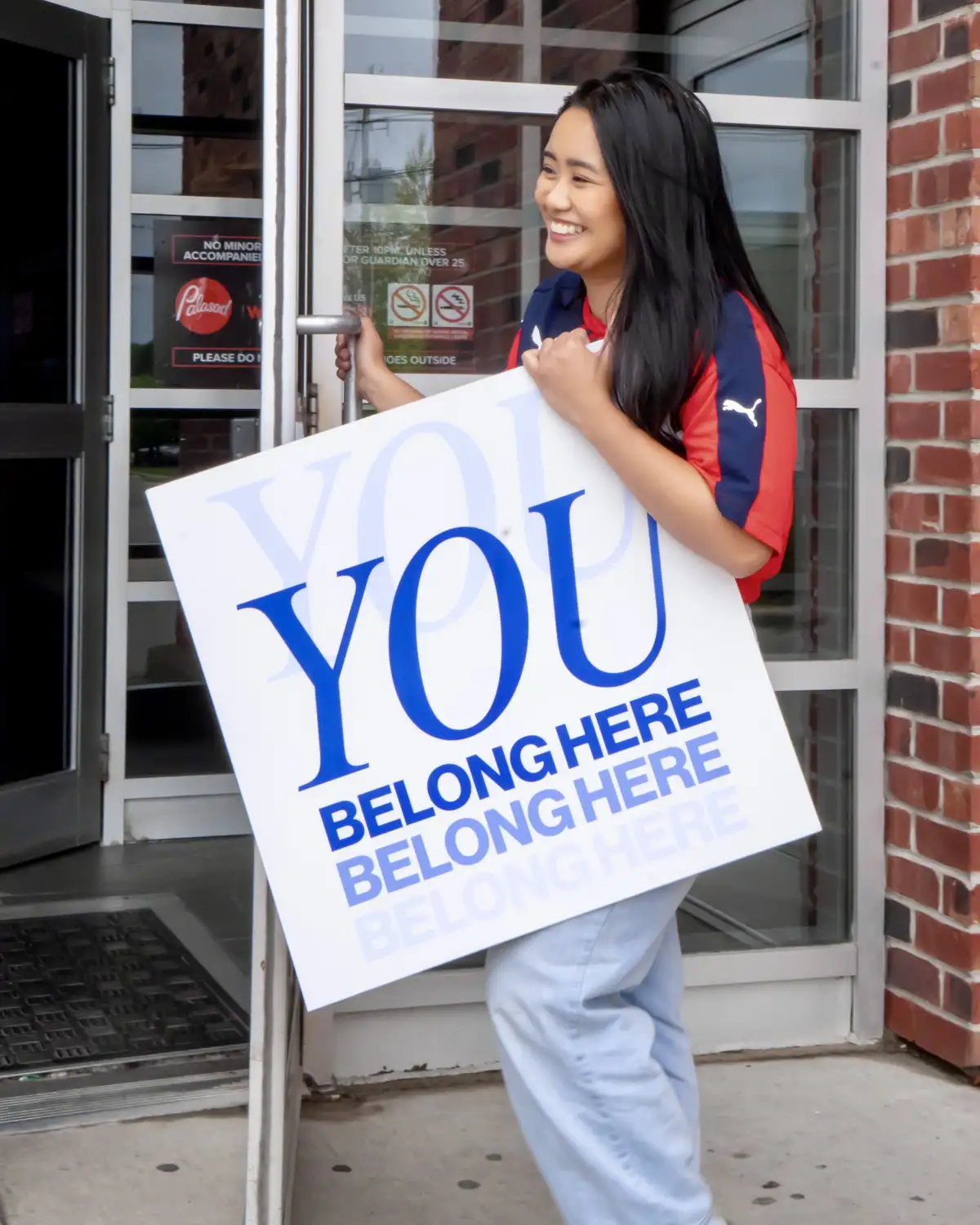 Smiling woman with long dark hair wearing a red and navy Puma shirt and light blue pants holding a sign that says 'YOU BELONG HERE' in blue letters while opening a glass door of a brick building.