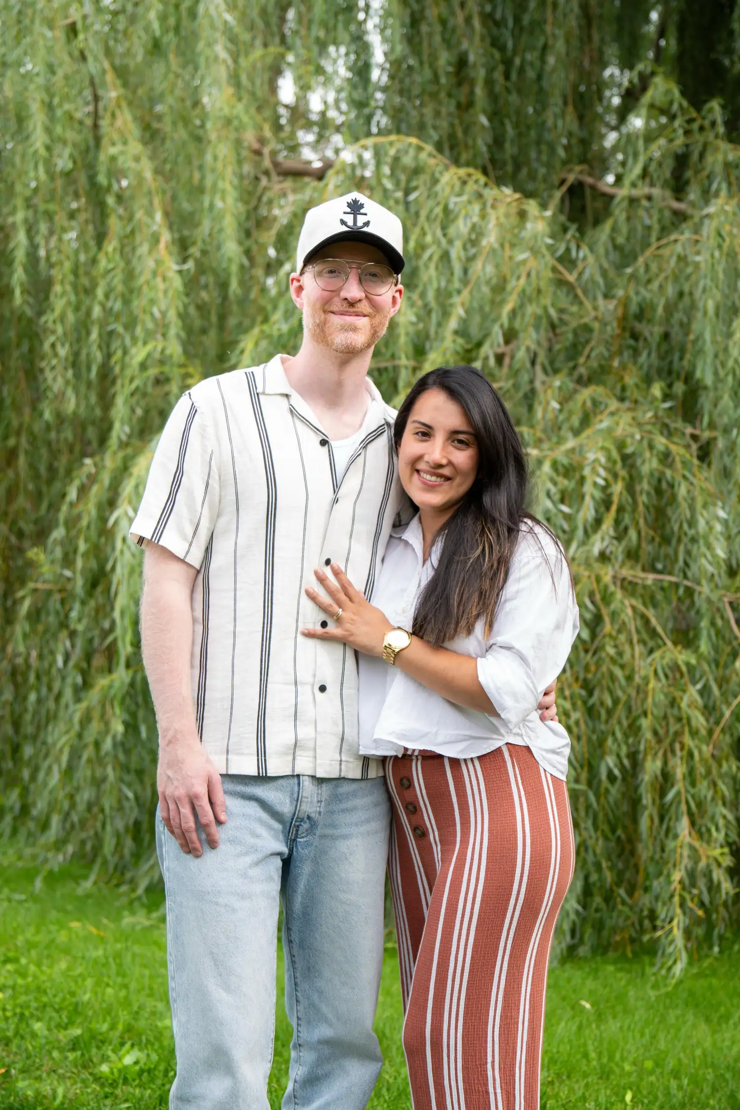 Smiling couple standing outdoors with greenery and hanging tree branches in the background. The man wears glasses, a white shirt with black stripes, light blue jeans, and a white hat with a black anchor and maple leaf emblem. The woman, standing close to him with her hand on his chest, wears a white blouse and rust-colored pants with vertical white stripes and buttons on the side.