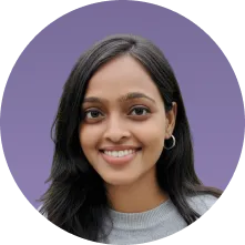 Smiling young woman with dark hair wearing hoop earrings and a gray top on a purple background.