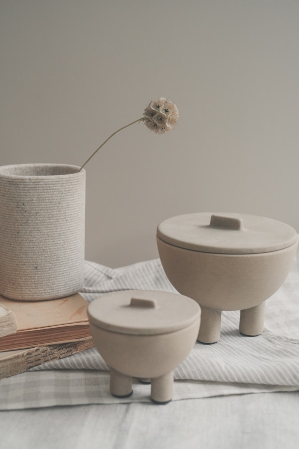 A serene still life composition featuring two sculptural ceramic containers with lids, a ribbed stone cup holding a single dried flower, and a stack of weathered books on soft striped fabric